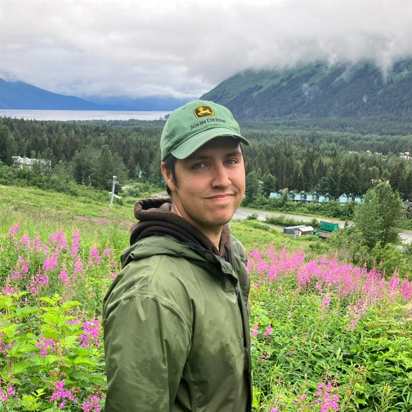 Young man in green jacket and cap standing in a field of pink flowers with mountains and cloudy sky in the background.