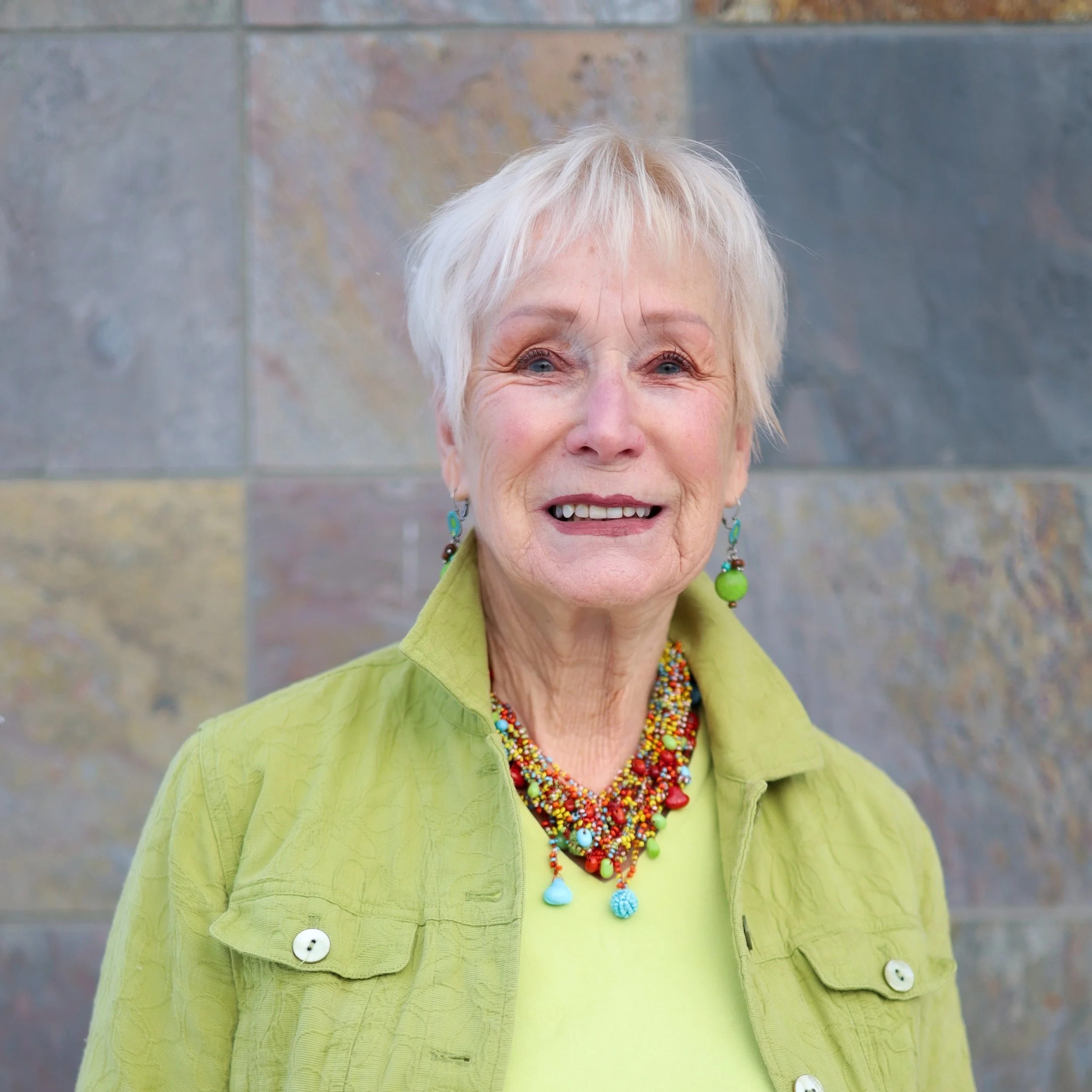 An elderly woman with short white hair and blue eyes, smiling, wearing a lime green shirt, colorful beaded jewelry, and earrings, standing in front of a textured stone wall.