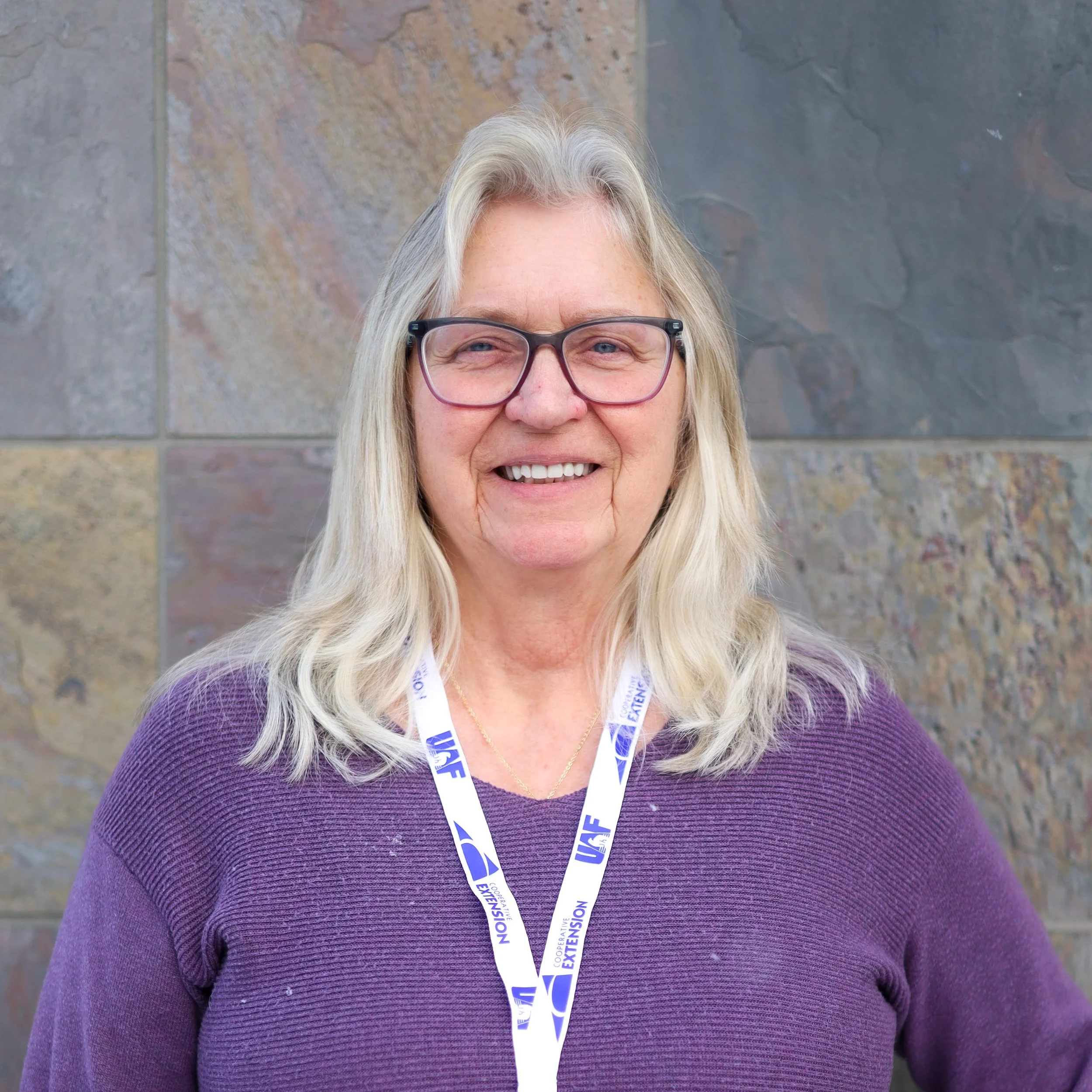 A smiling woman with glasses, wearing a purple sweater and a lanyard, standing outdoors against a stone wall.