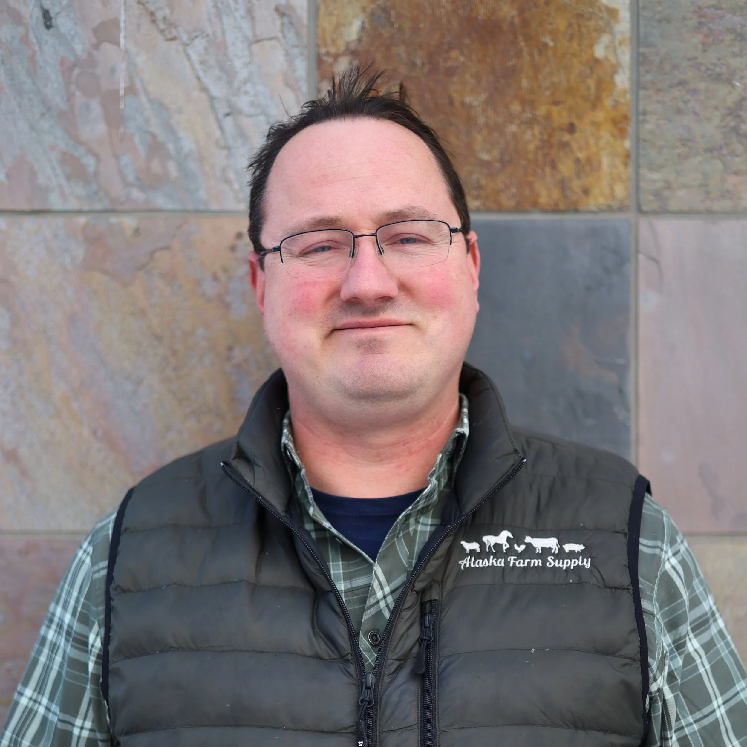 Man wearing glasses and a black puffer vest that has the logo and text, 'Alaska Farm Supply,' standing against a stone wall.