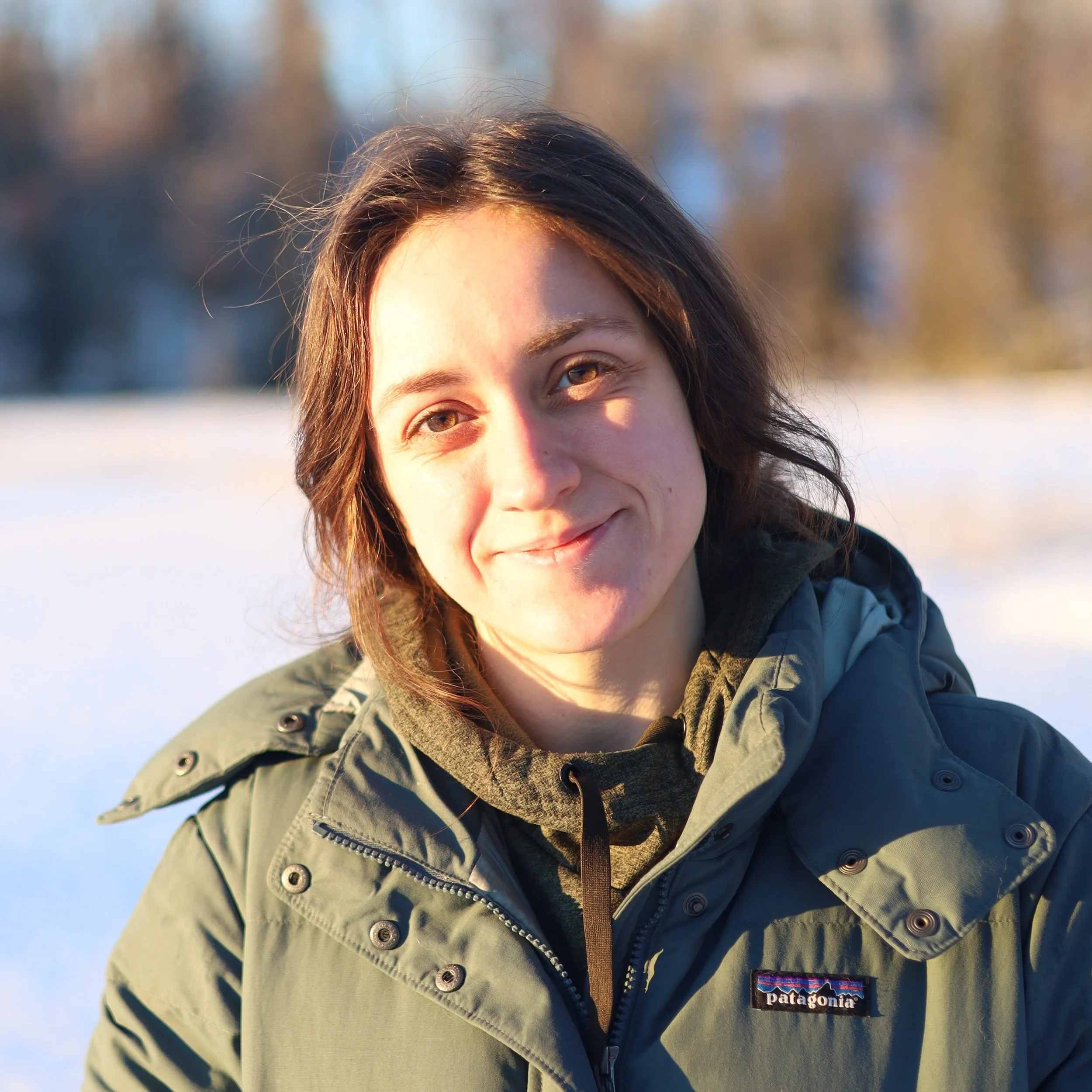 A woman with medium-length brown hair smiling outdoors in winter, wearing a green Patagonia jacket and a dark hoodie, with snow and trees in the background.