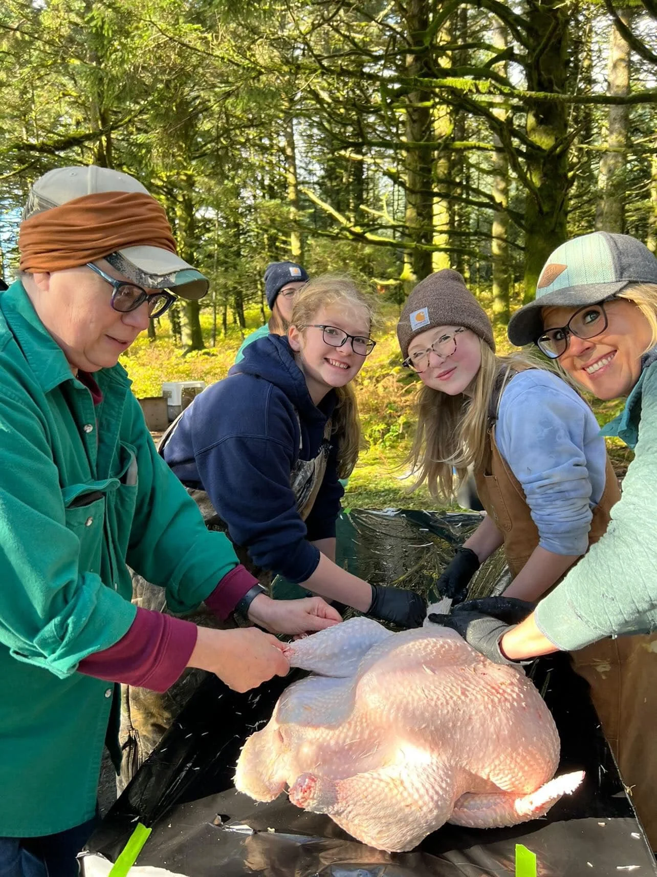 Group of people, including children and adults, preparing a large raw turkey outdoors in a forested area with trees and sunlight.