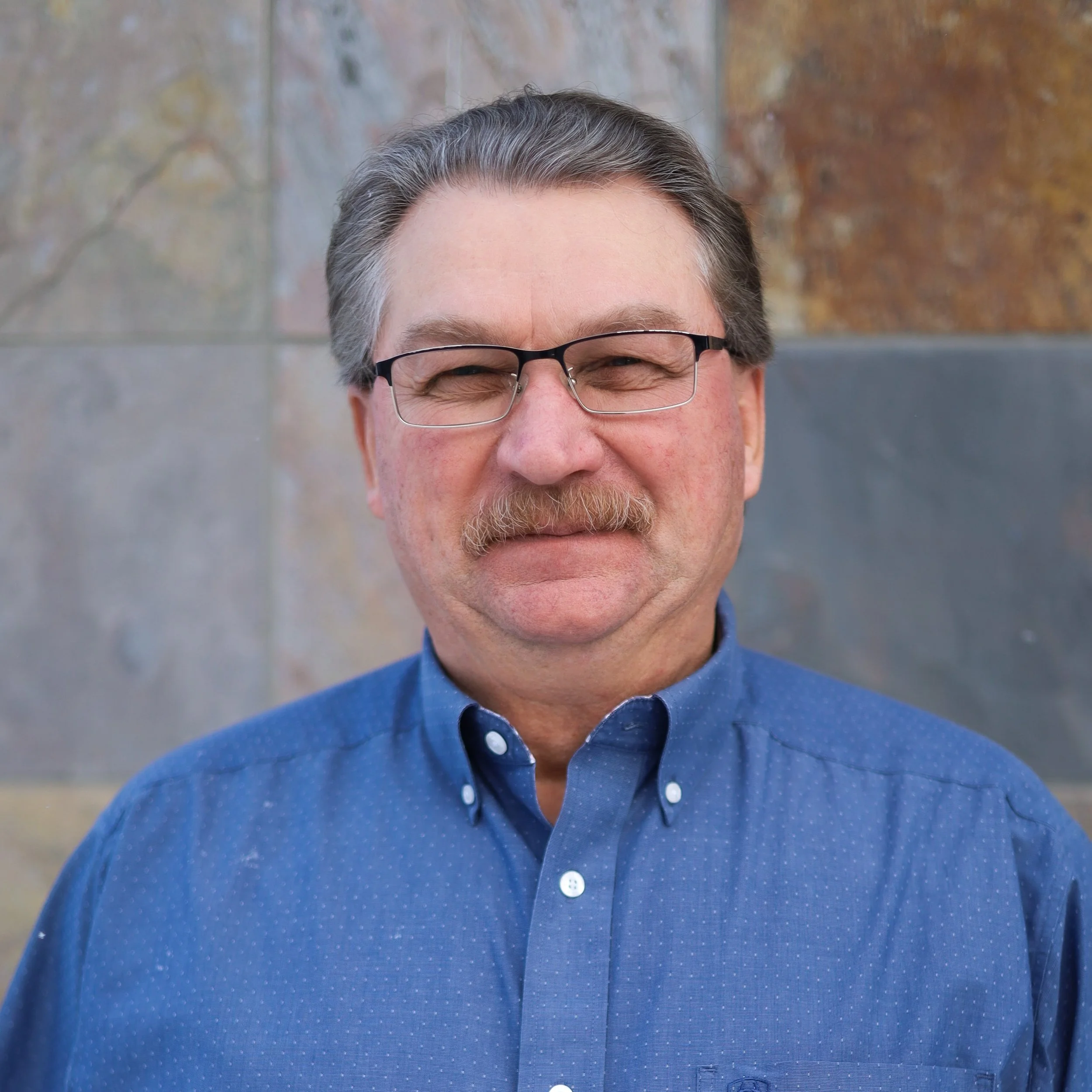 A middle-aged man with gray hair, glasses, and a mustache, wearing a blue button-up shirt, standing outdoors in front of a multicolored stone wall.