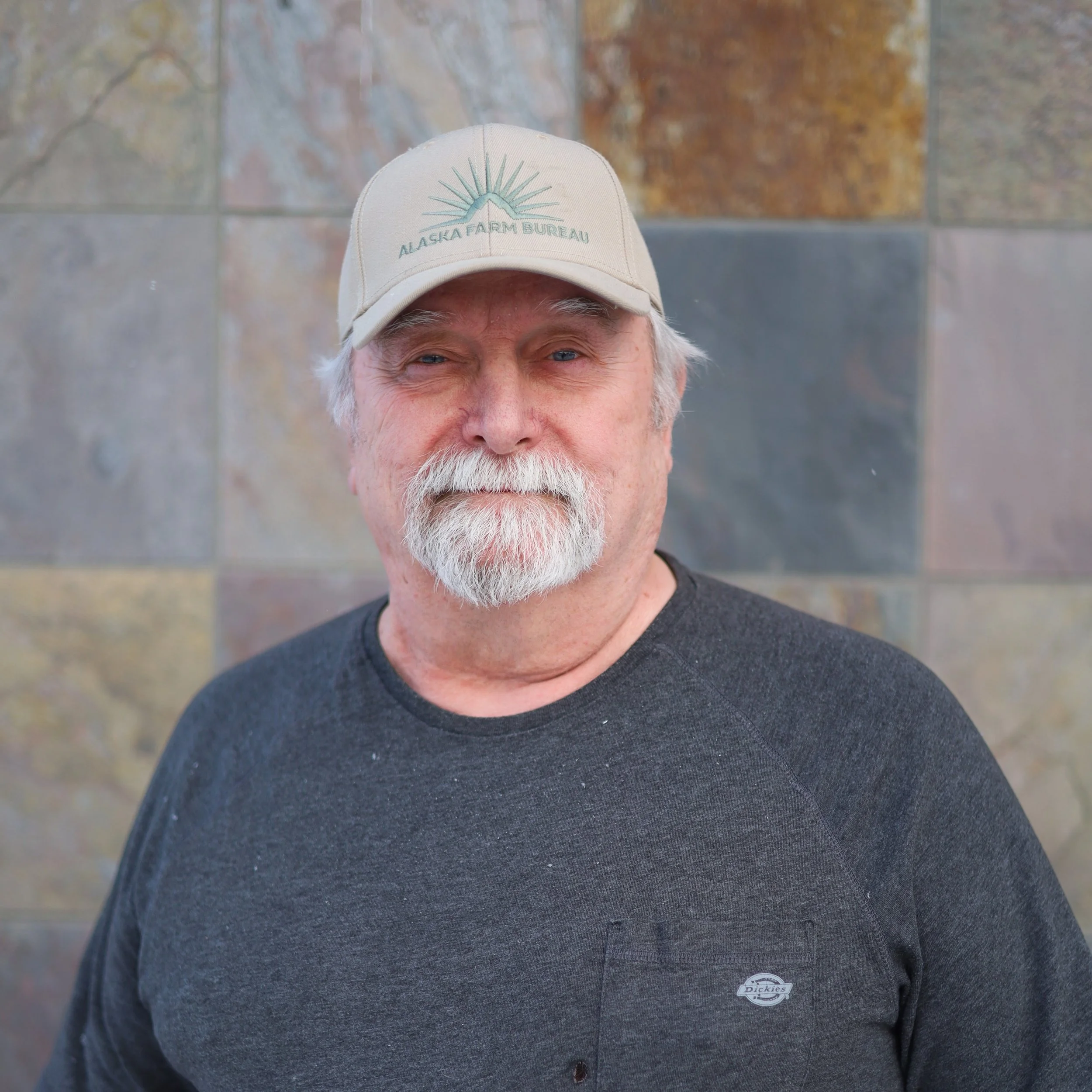 An older man with a white beard and mustache, wearing a tan baseball cap with 'Alaska Farm Bureau' logo, and a dark gray shirt, standing in front of a stone wall.