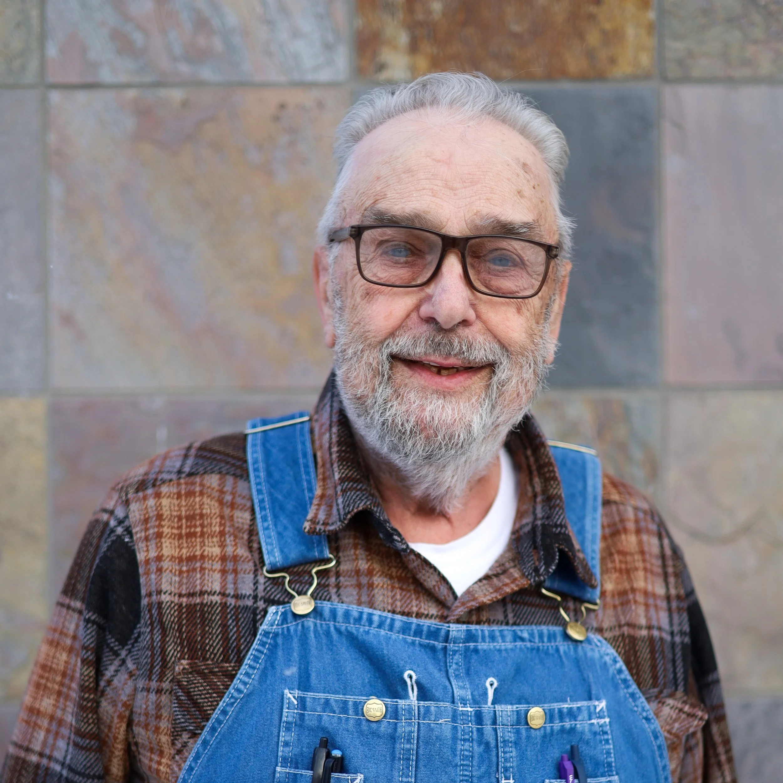 Elderly man with gray hair, glasses, and a beard smiling, wearing a plaid shirt and denim apron, standing in front of a brick wall.
