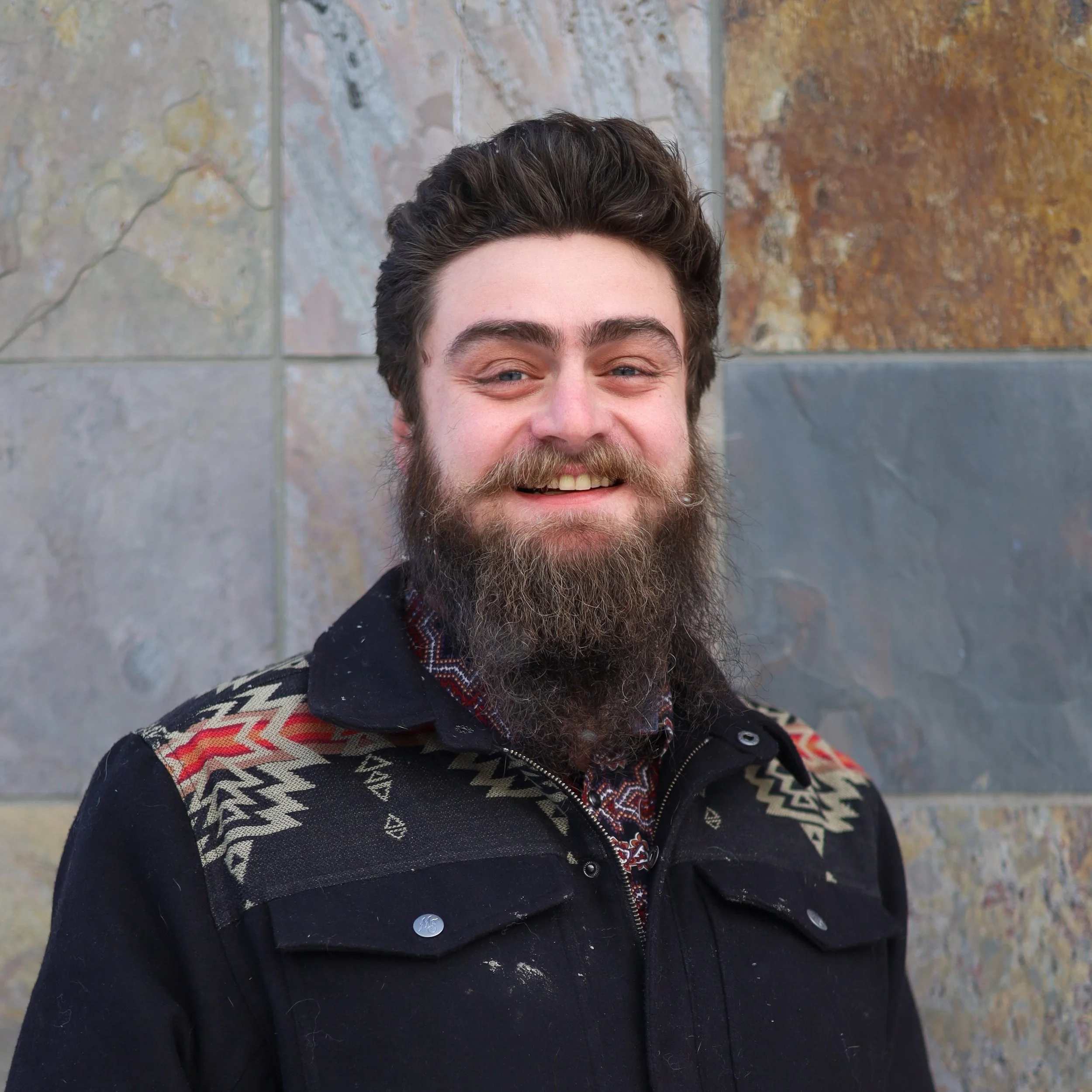 A smiling man with a full beard and mustache, wearing a black jacket with colorful patterns on the shoulders, standing in front of a stone wall.