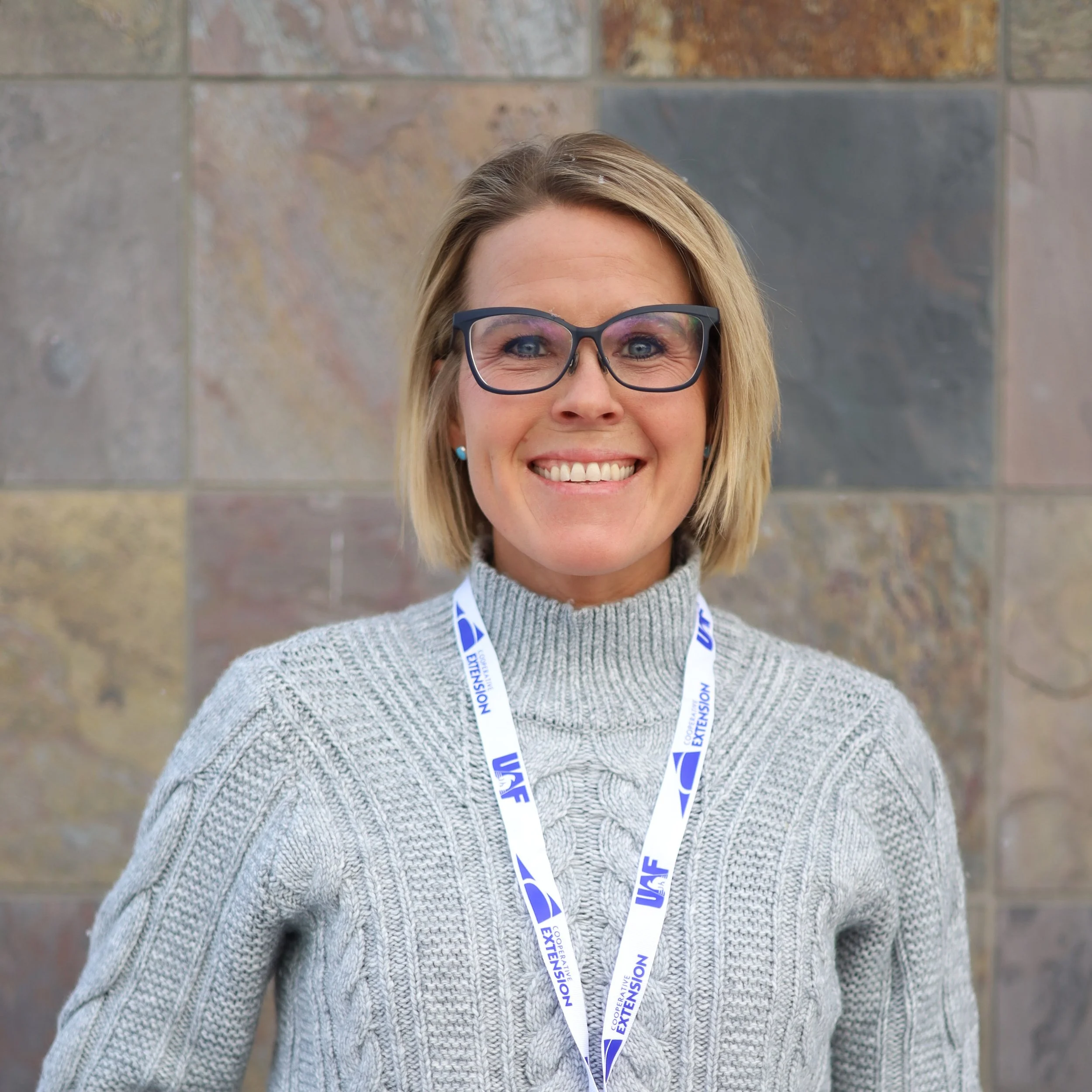 A woman with shoulder-length blonde hair, wearing glasses, a gray cable-knit sweater, and a lanyard with a blue and white logo, smiling in front of a stone wall.