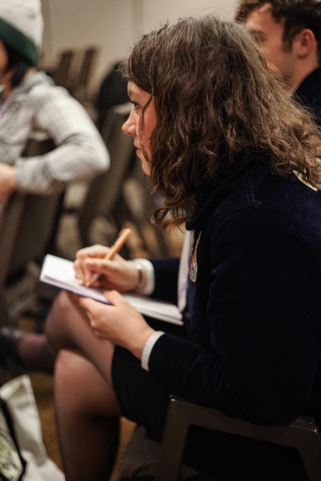 A woman with curly brown hair sitting and taking notes during a meeting or conference.