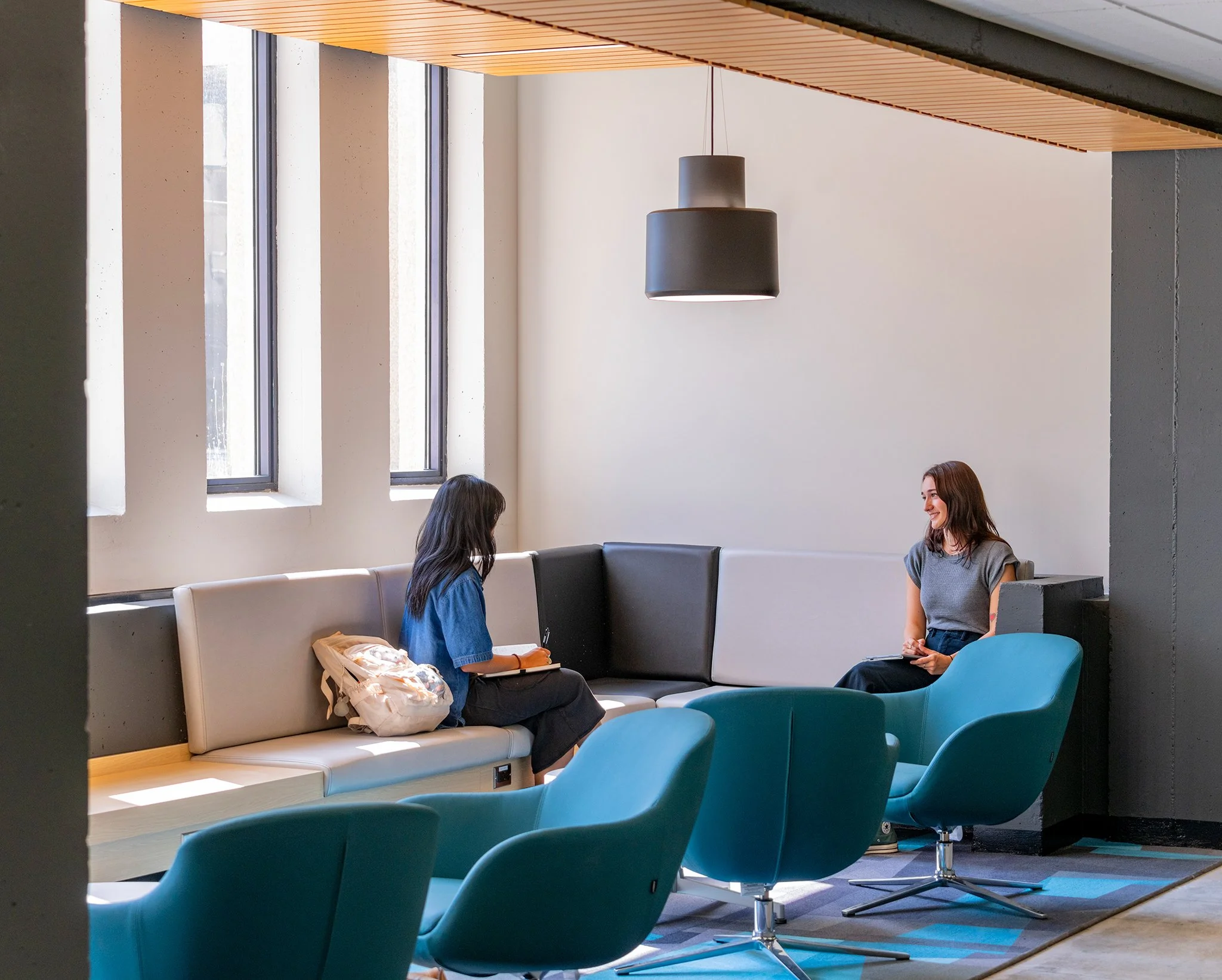 Two women sitting and talking in a modern, brightly lit lounge area with large windows, teal lounge chairs, and black and white seating.