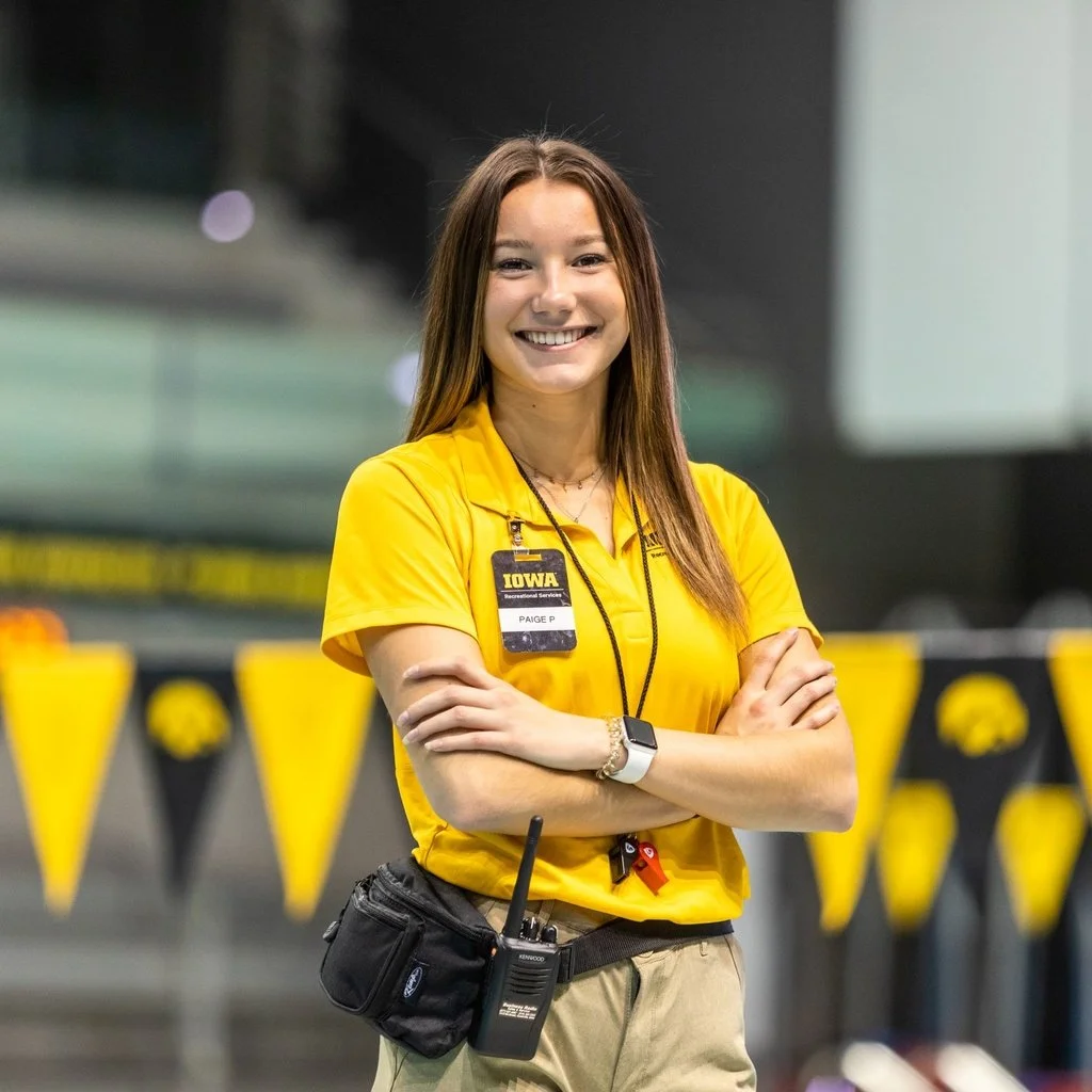 A young woman in a yellow uniform with an ID badge that says "IOWA" and "PAIGE P" stands smiling with arms crossed inside a facility with black and yellow decor.