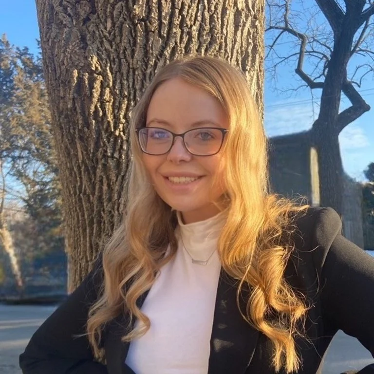 A young woman with long blonde hair, glasses, and wearing a black blazer and white top, smiling outdoors in front of a large tree with a rough textured trunk, during daytime with clear blue sky.