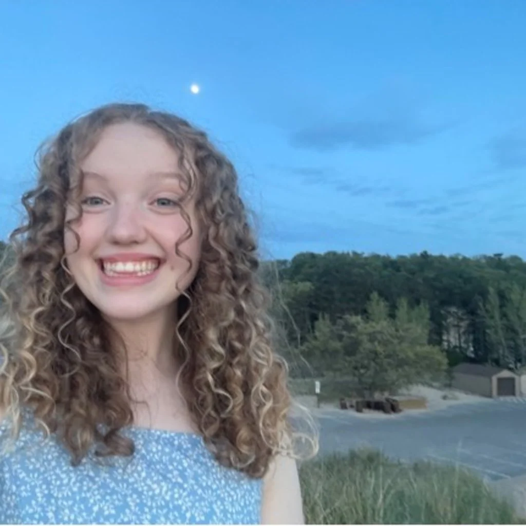 A young woman with curly hair taking a selfie outdoors during dusk, with a bright moon and cloudy sky in the background.