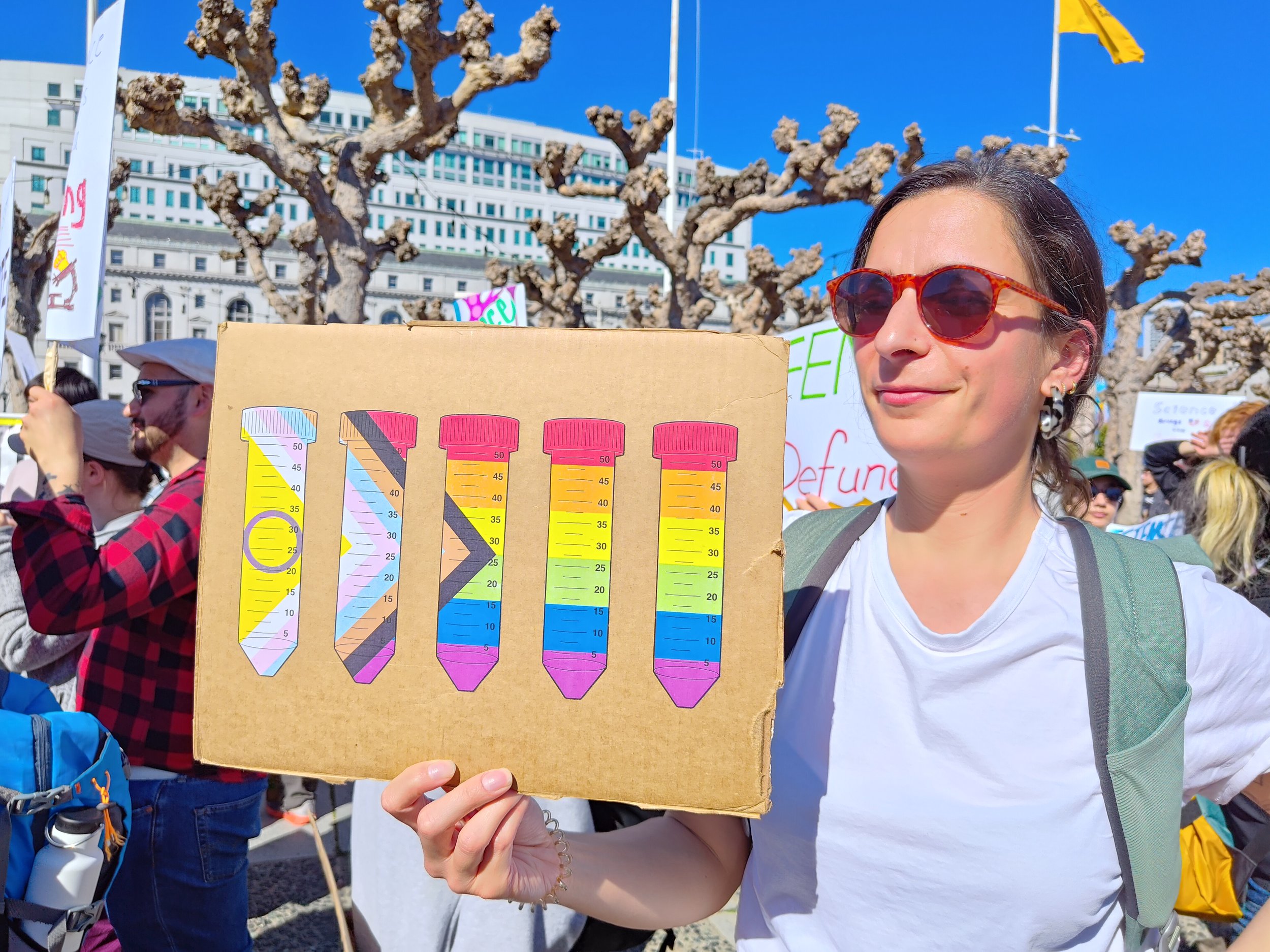 Protester with sign depicting a row of test tubes filled in with the Progress Pride flag.