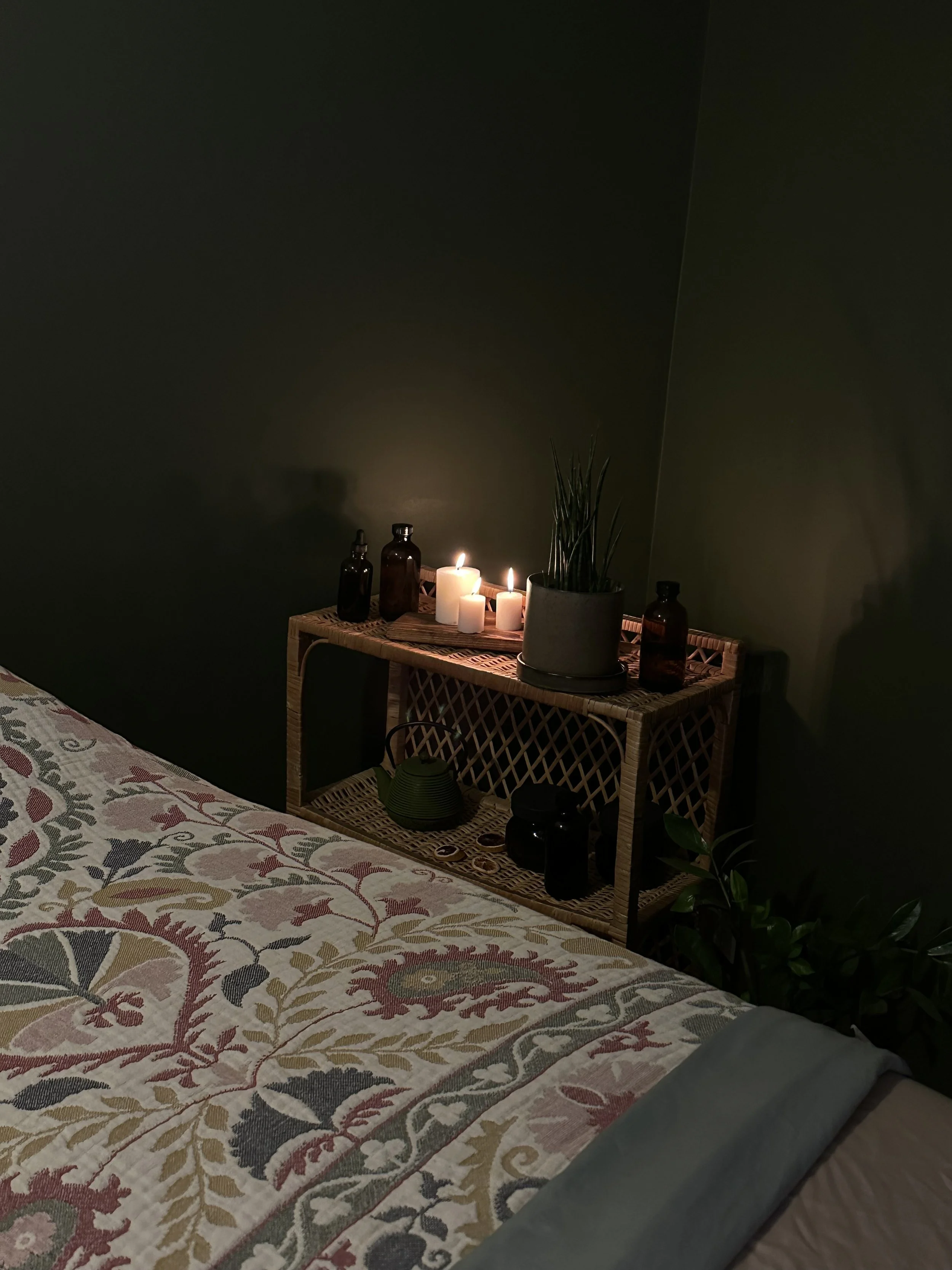 A bedroom with a bedside table holding candles, bottles, a plant, and a teapot, with a bed covered in a floral quilt.