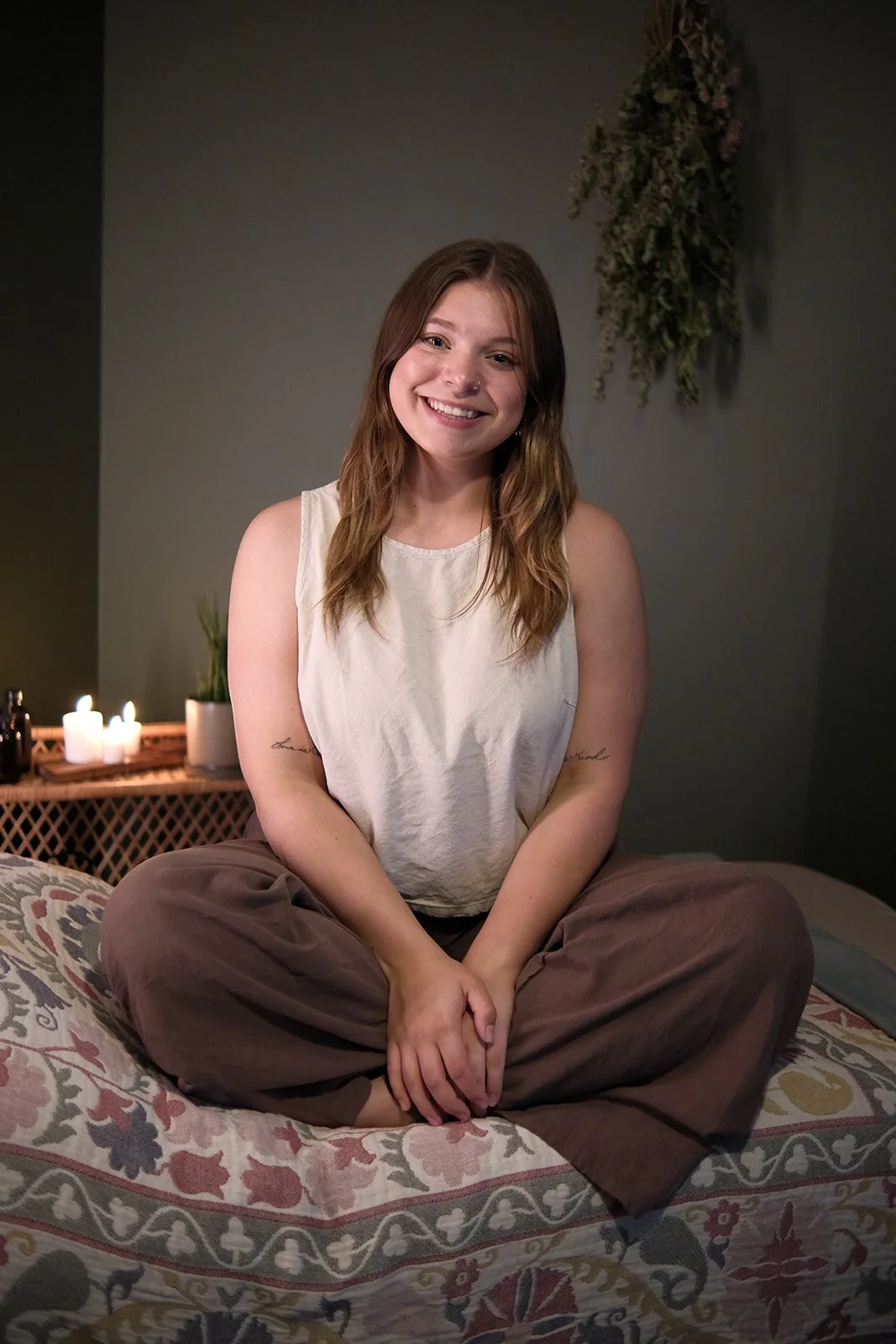 Young woman sitting cross-legged on a bed in a dimly lit room with candles, a plant, and a wall hanging in the background, smiling at the camera.