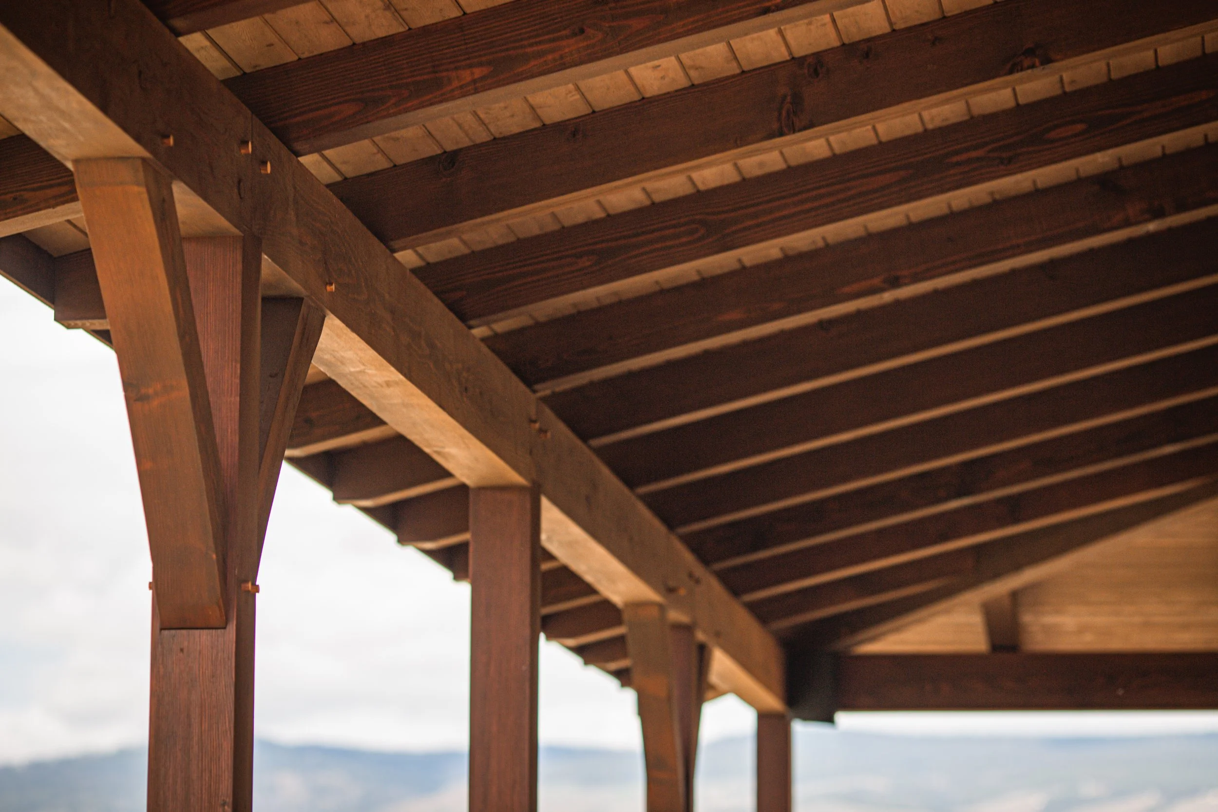Close-up view of a wooden pergola roof structure with beams and joists, with a cloudy sky and mountains in the background.