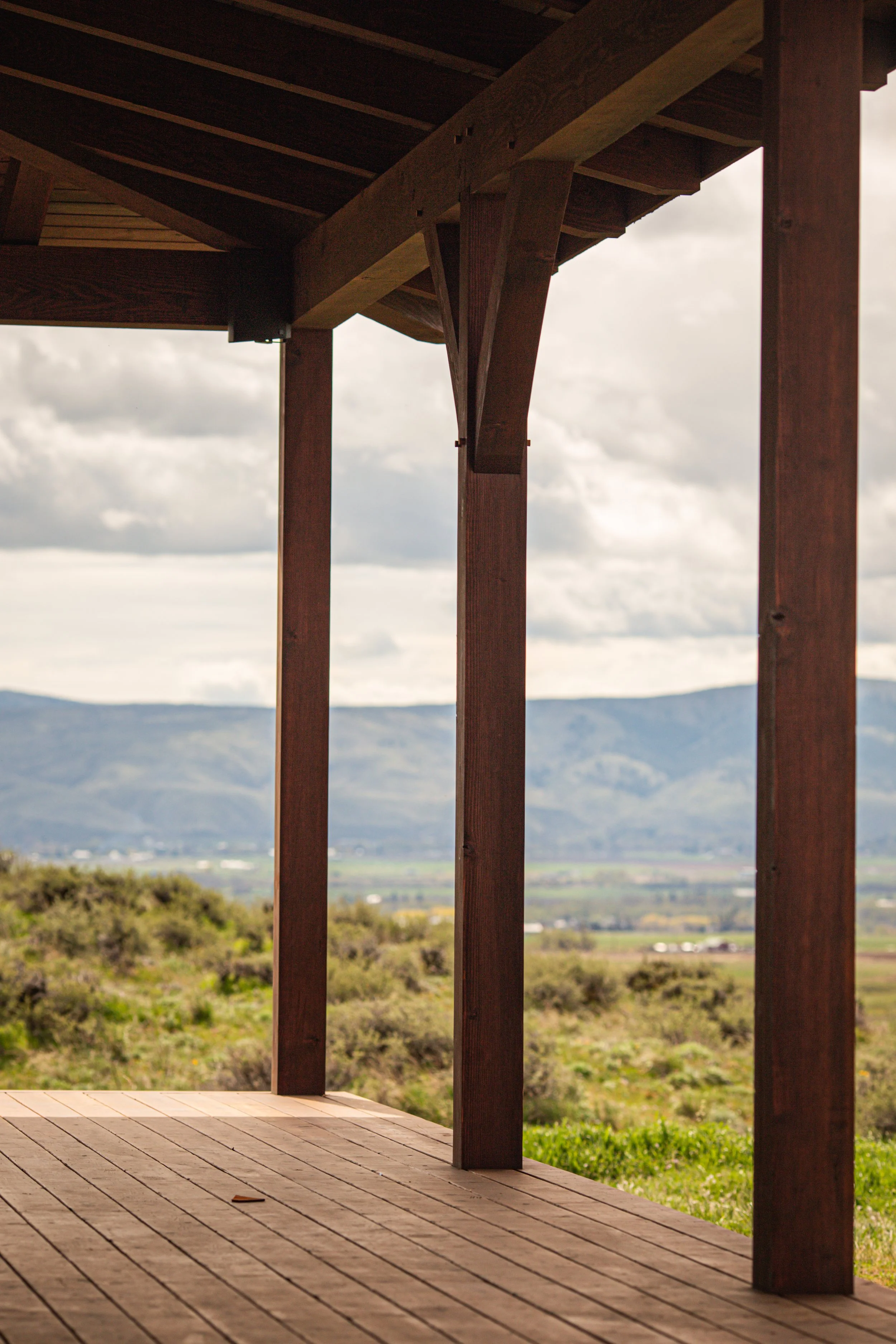 View from a wooden deck with vertical wooden beams, overlooking green fields and distant mountains under a cloudy sky.