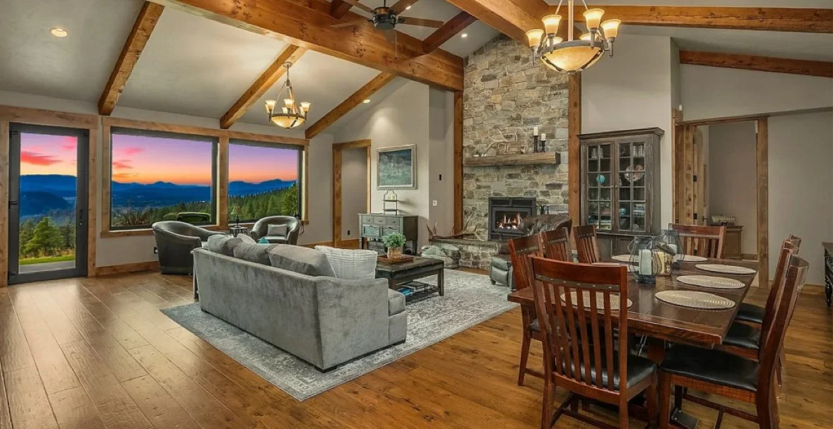 Living room with hardwood floors, a large gray sofa, two leather armchairs, a stone fireplace, a wooden dining table for six, and large windows with a mountain view at sunset.