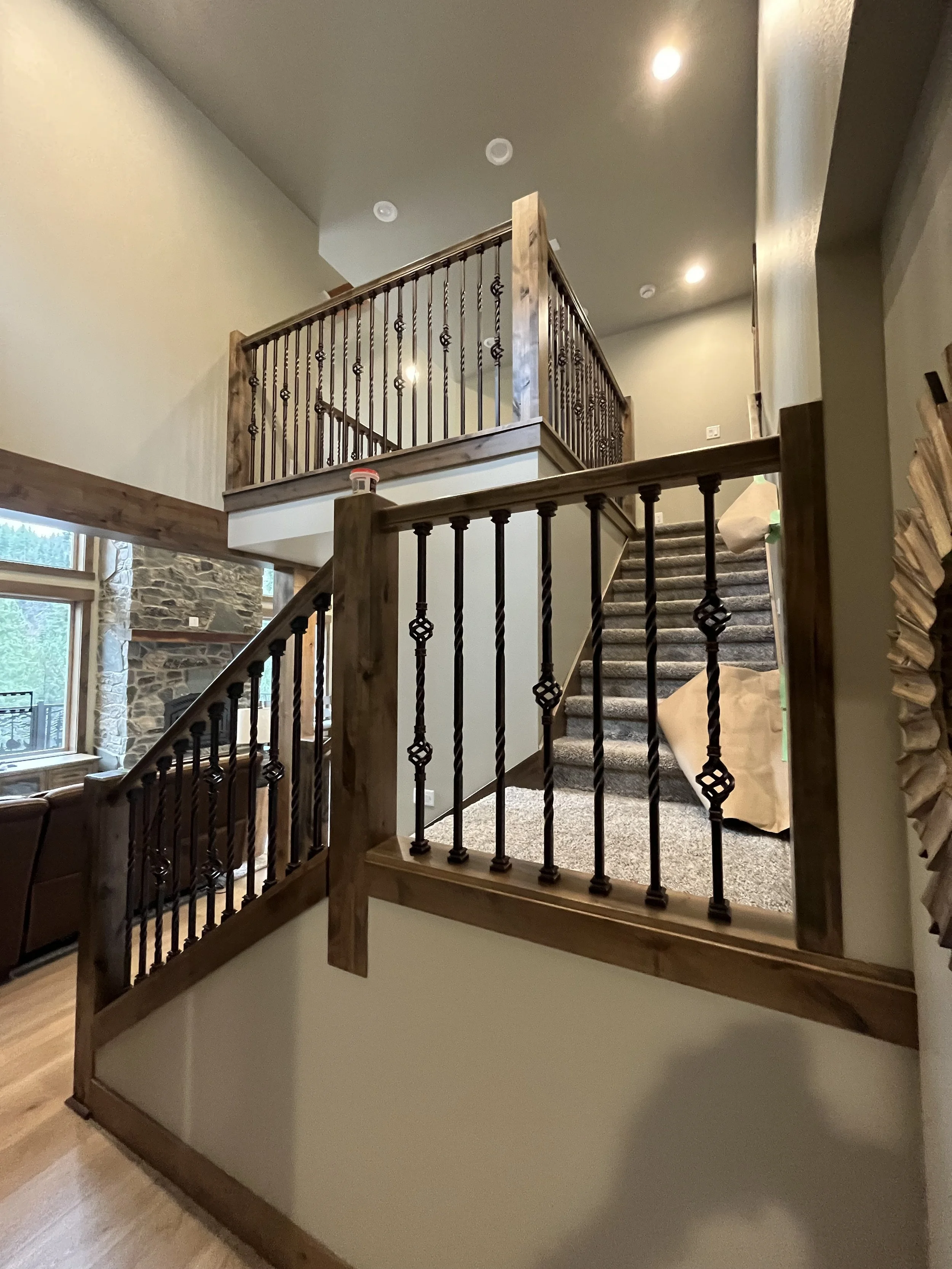 Interior view of a staircase with wooden and wrought iron railing, carpeted stairs, large stone fireplace, and windows showing trees outside.