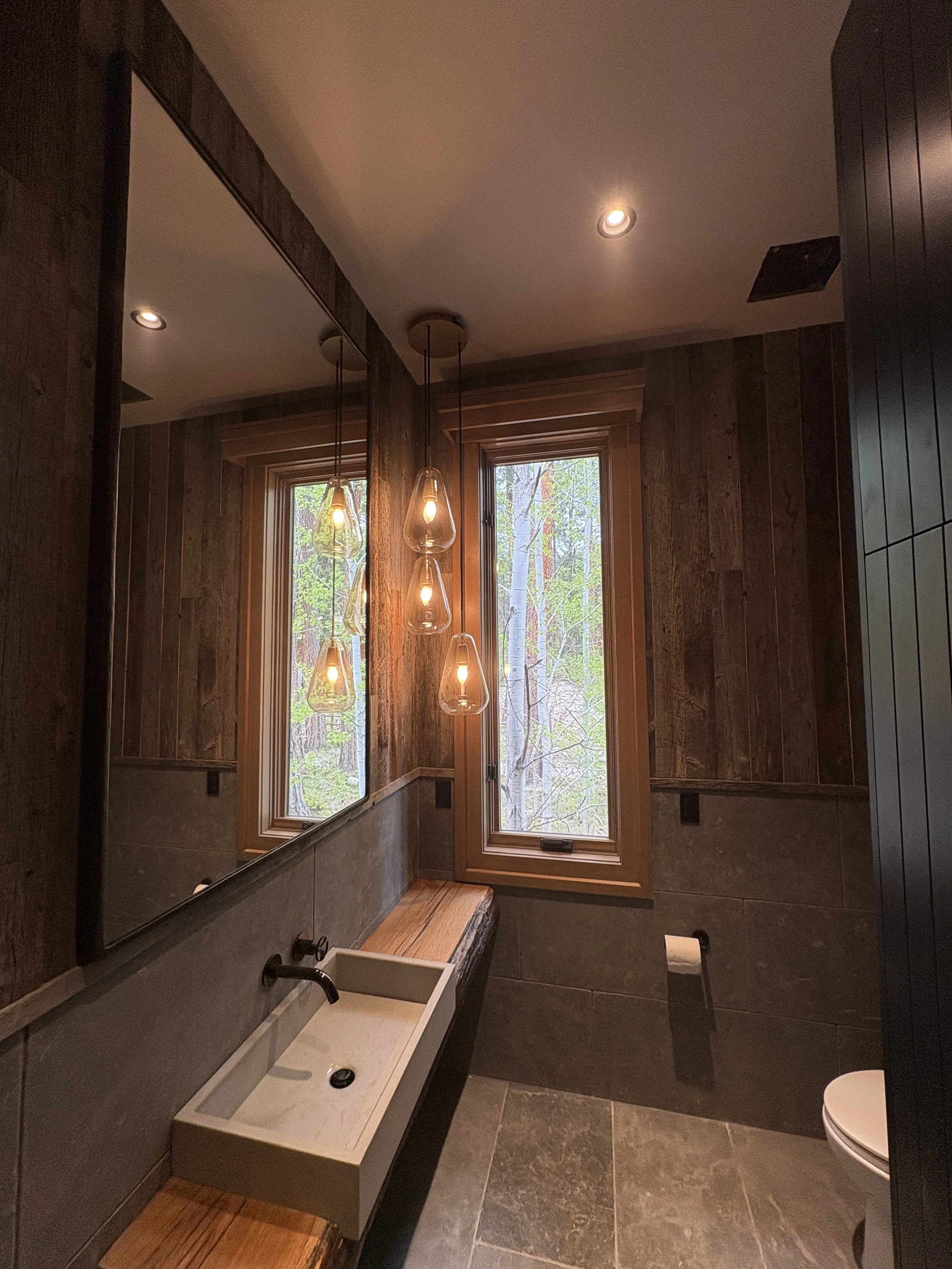 A rustic-style bathroom with wooden walls and modern fixtures. There is a rectangular vessel sink with a black faucet, a toilet on the right, a mirror on the left, and a large window showing trees outside. Multiple pendant lights hang from the ceiling, illuminating the space.