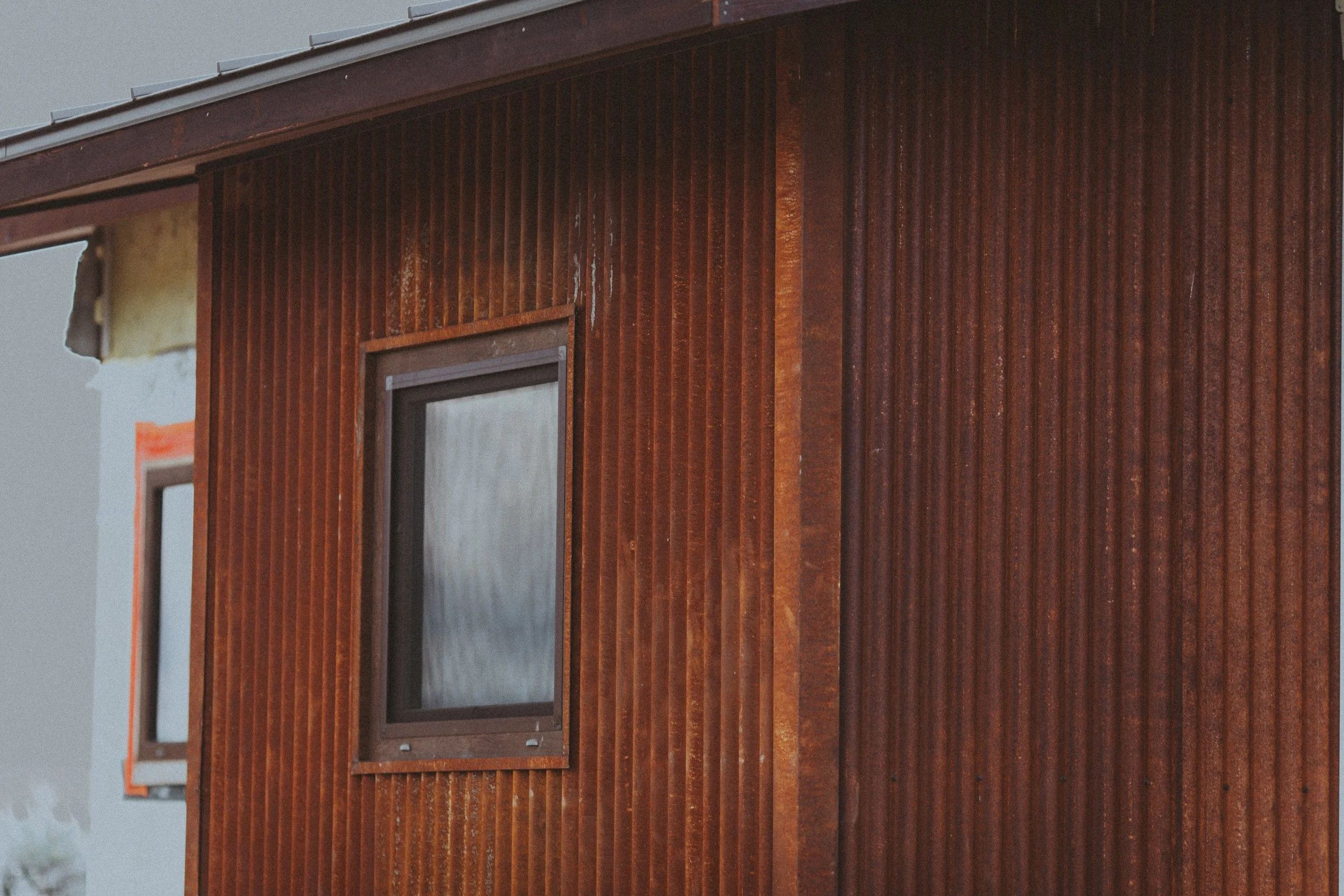 Close-up of a building's exterior wall covered with vertical rusted metal siding and a window with a wooden frame.
