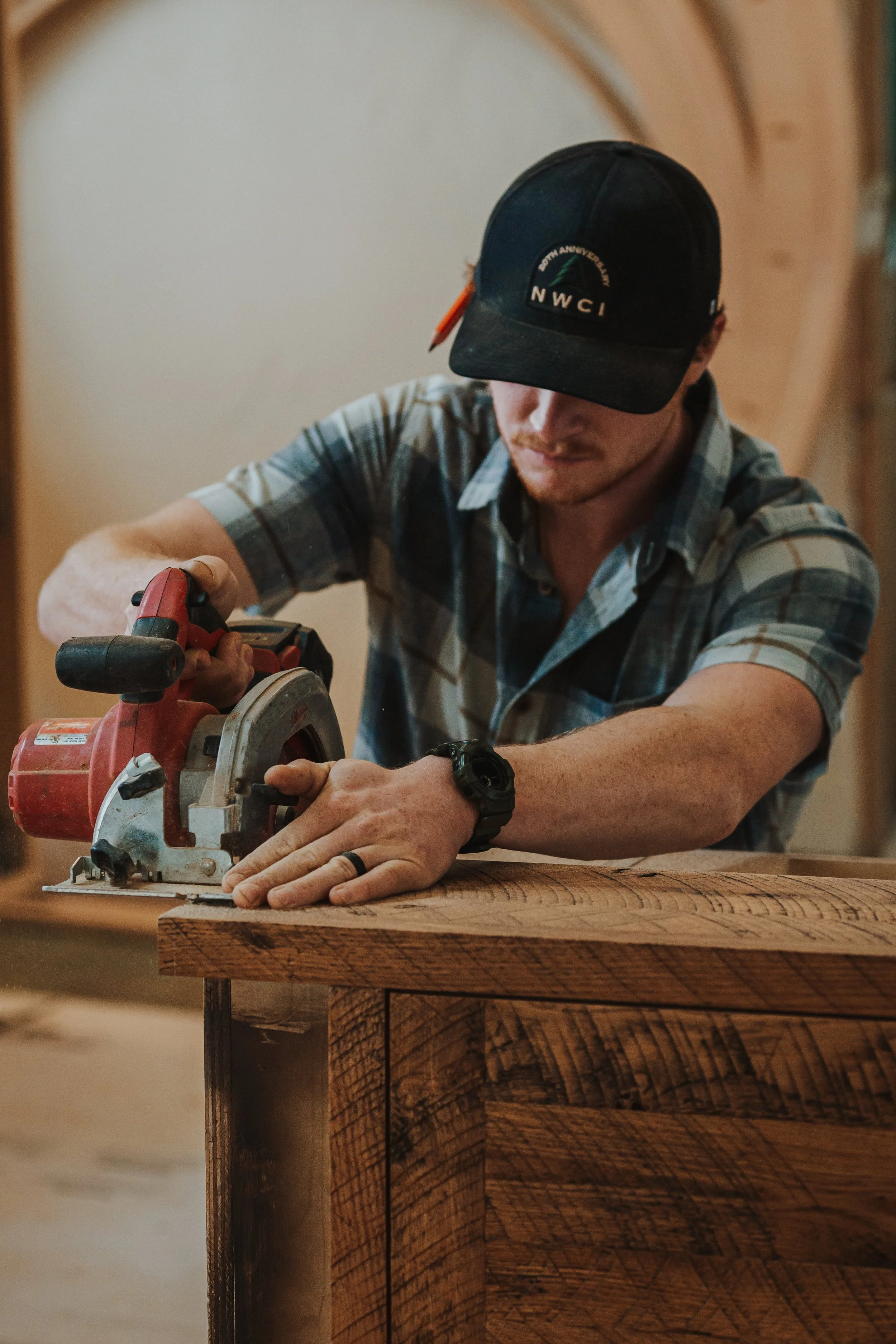 A man wearing a black cap and a plaid shirt is using a circular saw to cut a piece of wood on a woodworking table.