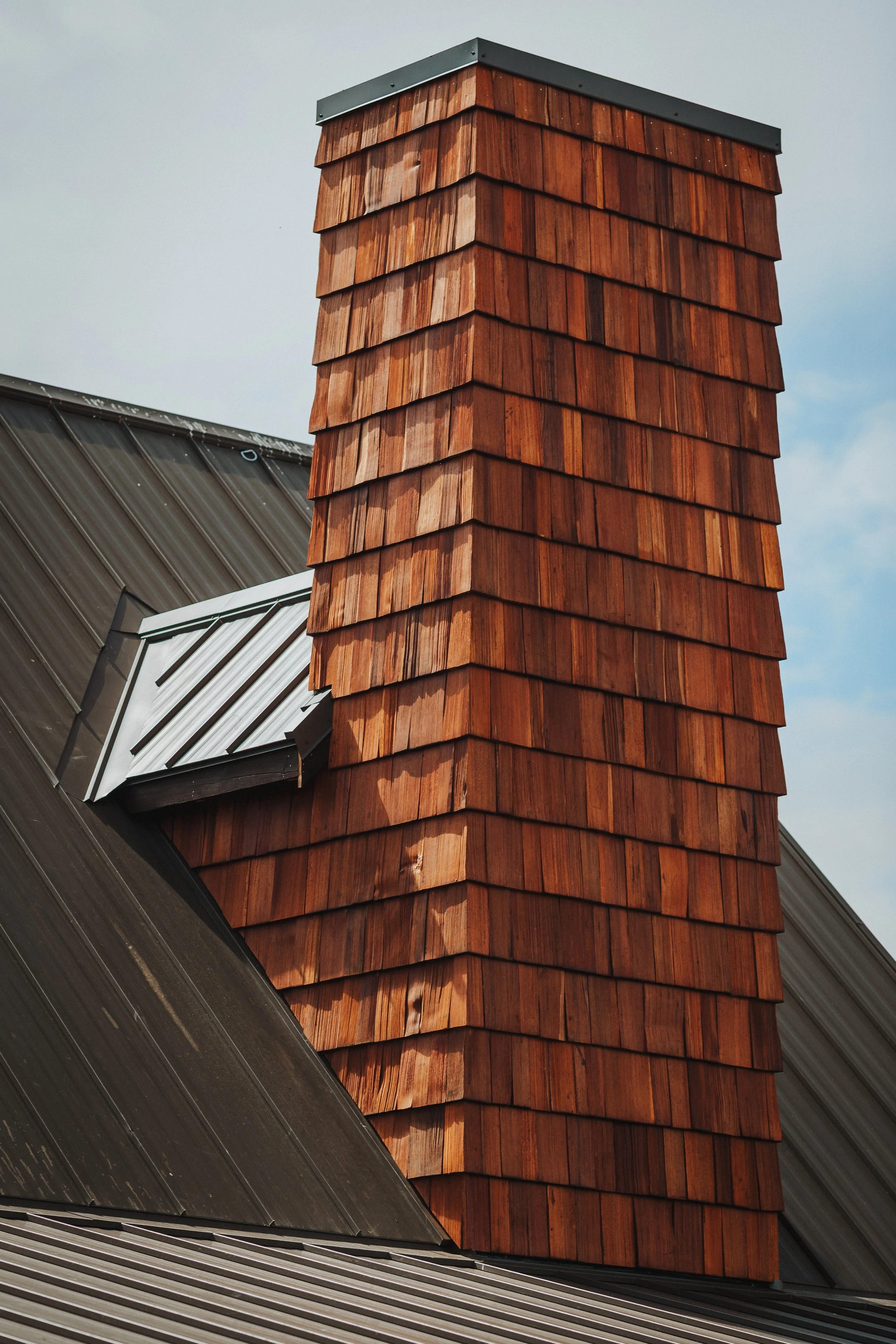 A large chimney covered with reddish-brown wooden shingles on a roof made of metal panels.