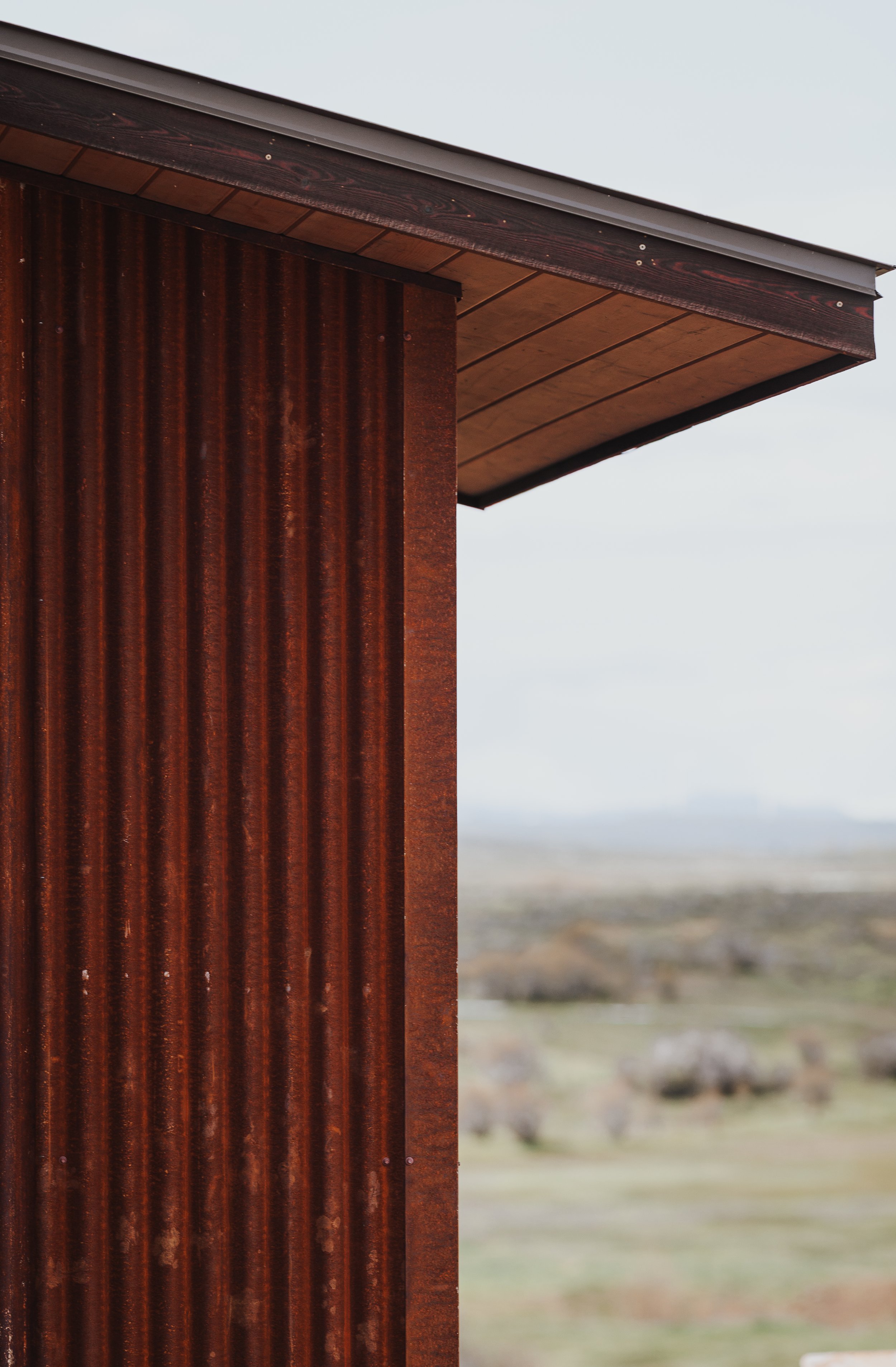 Close-up of a rusty metal building corner with a brown wooden roof overhang, against a blurred outdoor landscape background.