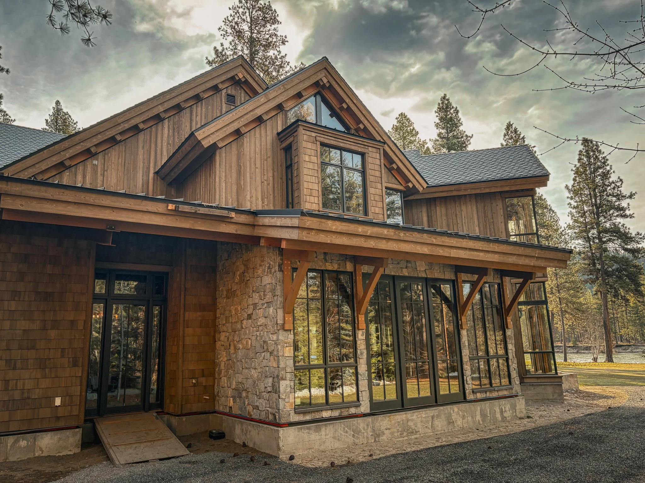 A house under construction with wood and stone exterior, large windows, and a gabled roof, surrounded by trees and a cloudy sky.