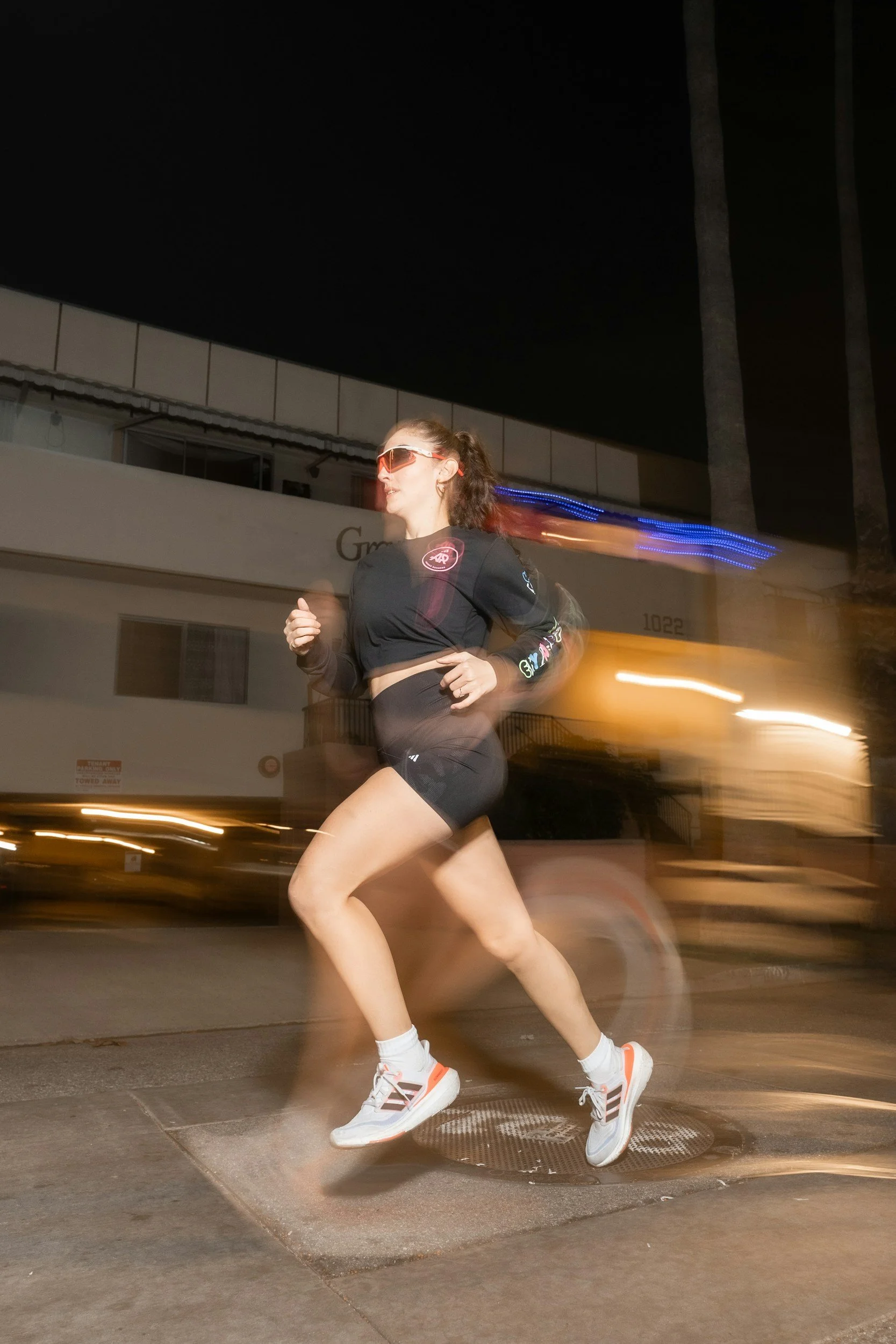 A woman jogging at night on a city street wearing sunglasses, a black athletic outfit, and running shoes, with light trails behind her indicating motion.