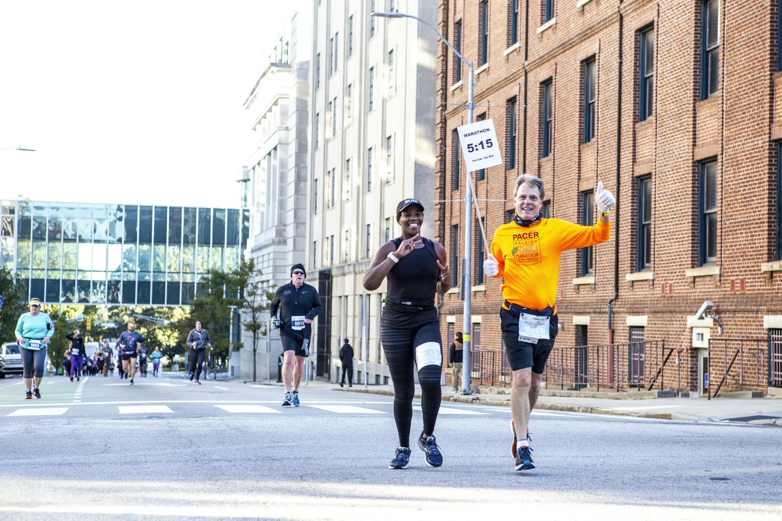 Marathon runners crossing the finish line on a city street, with a male runner in a bright orange shirt and a female runner in black in the foreground, and other runners behind them.