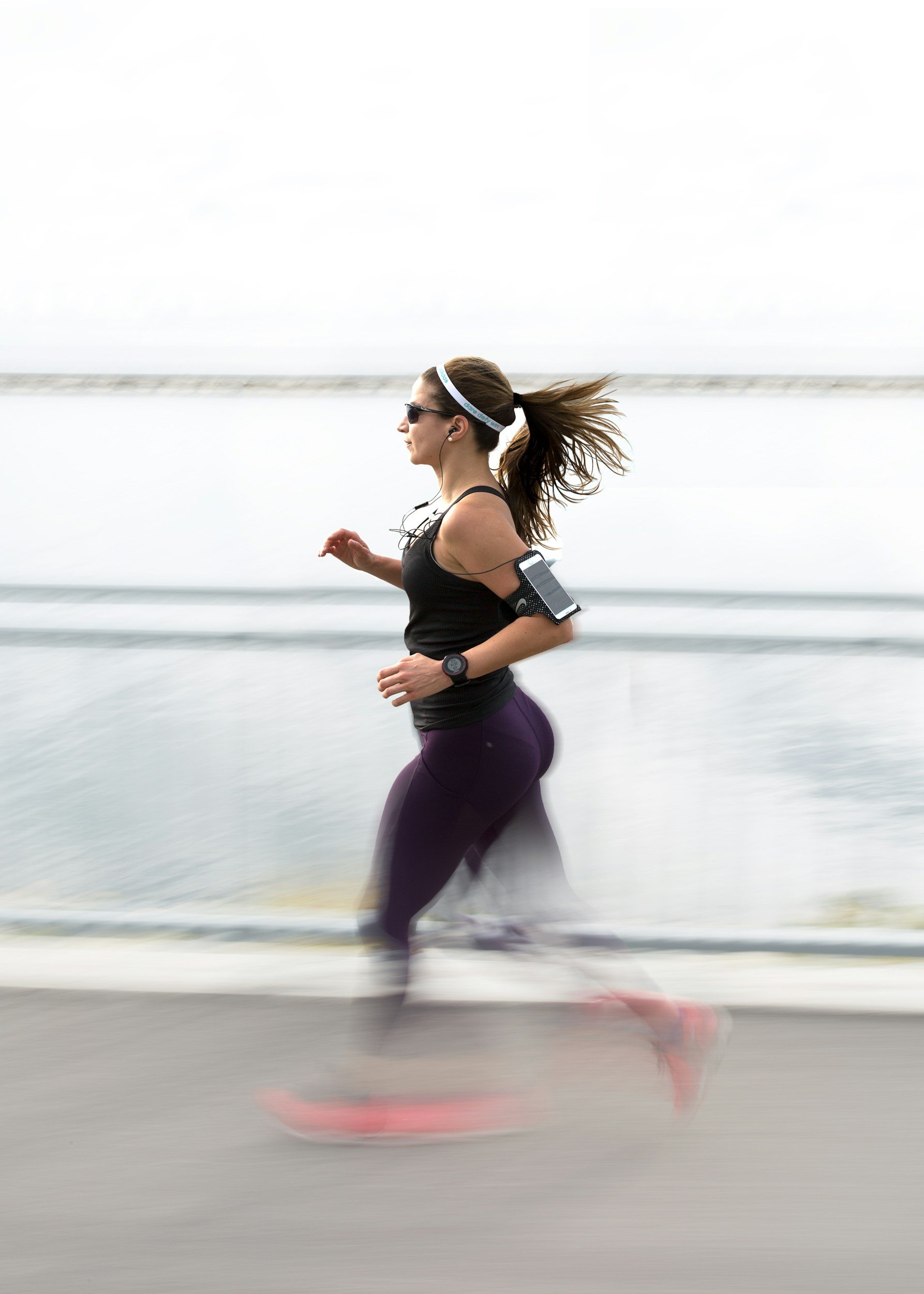 A woman jogging along a beachside road with the ocean in the background. She is wearing sunglasses, a black tank top, purple leggings, and earphones, with her hair pulled back and flowing behind her.