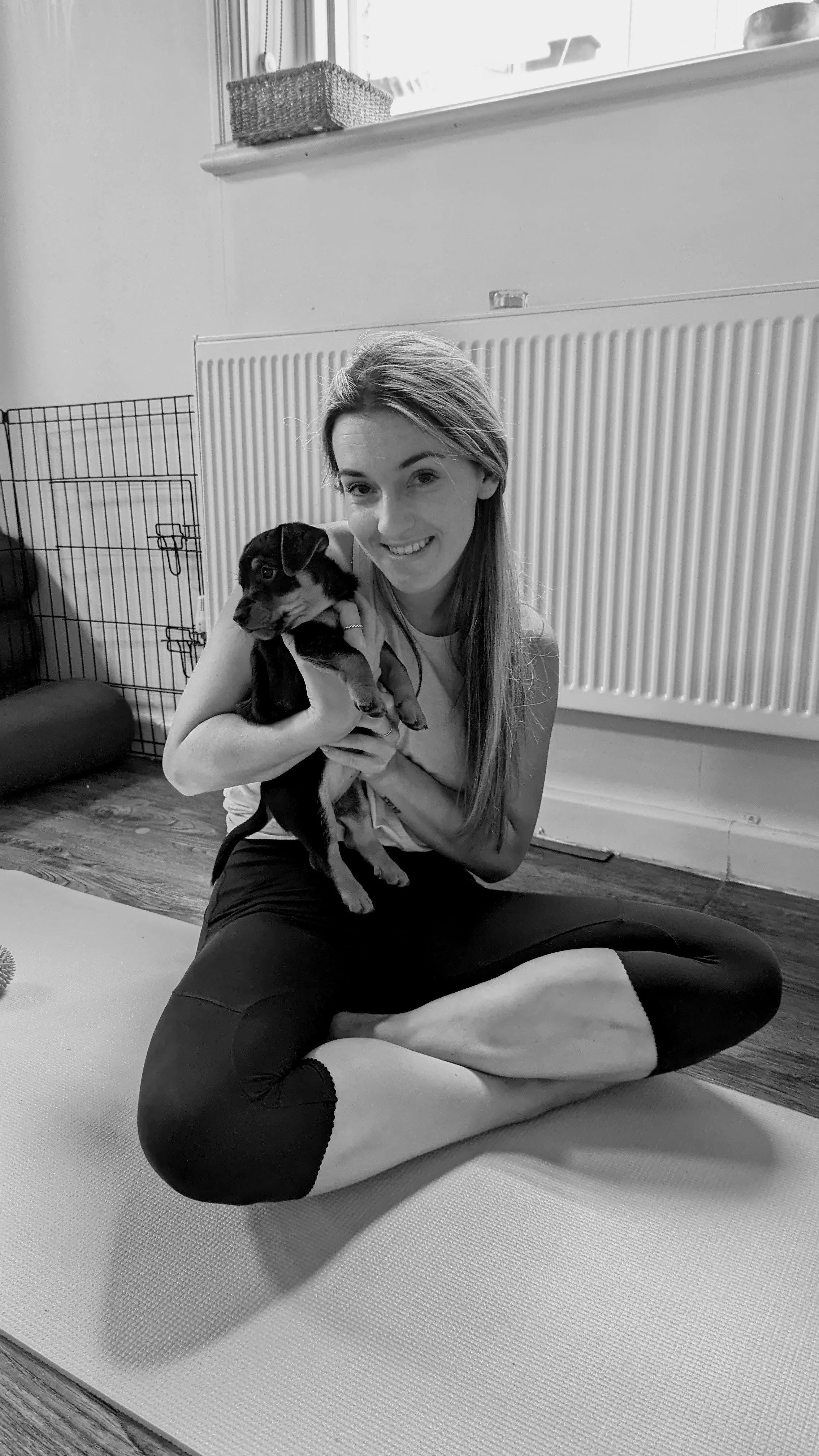 A smiling woman with long hair sitting cross-legged on a yoga mat, holding a small black and white puppy. The room has wooden flooring, a radiator, and a wire dog crate in the background.