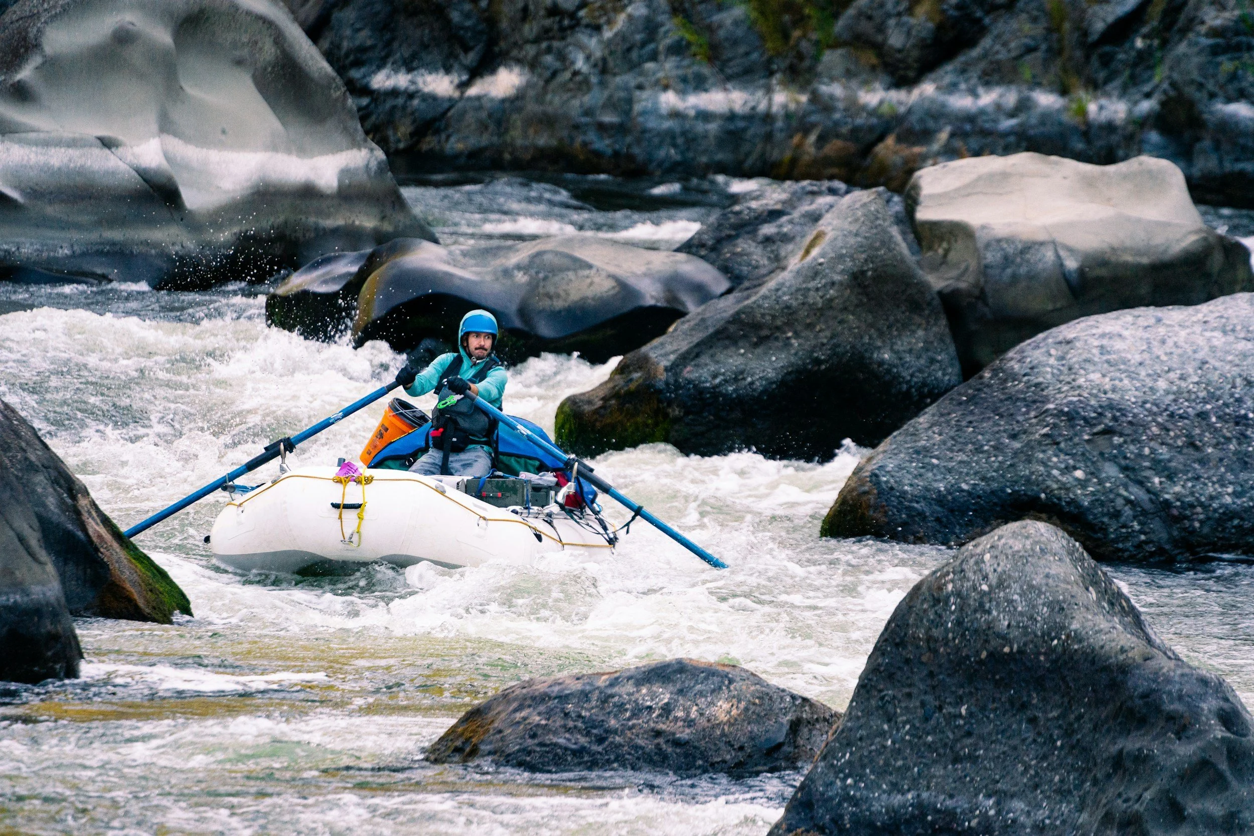 A person wearing a blue helmet and life vest paddling a white inflatable raft through rushing river water surrounded by large rocks.