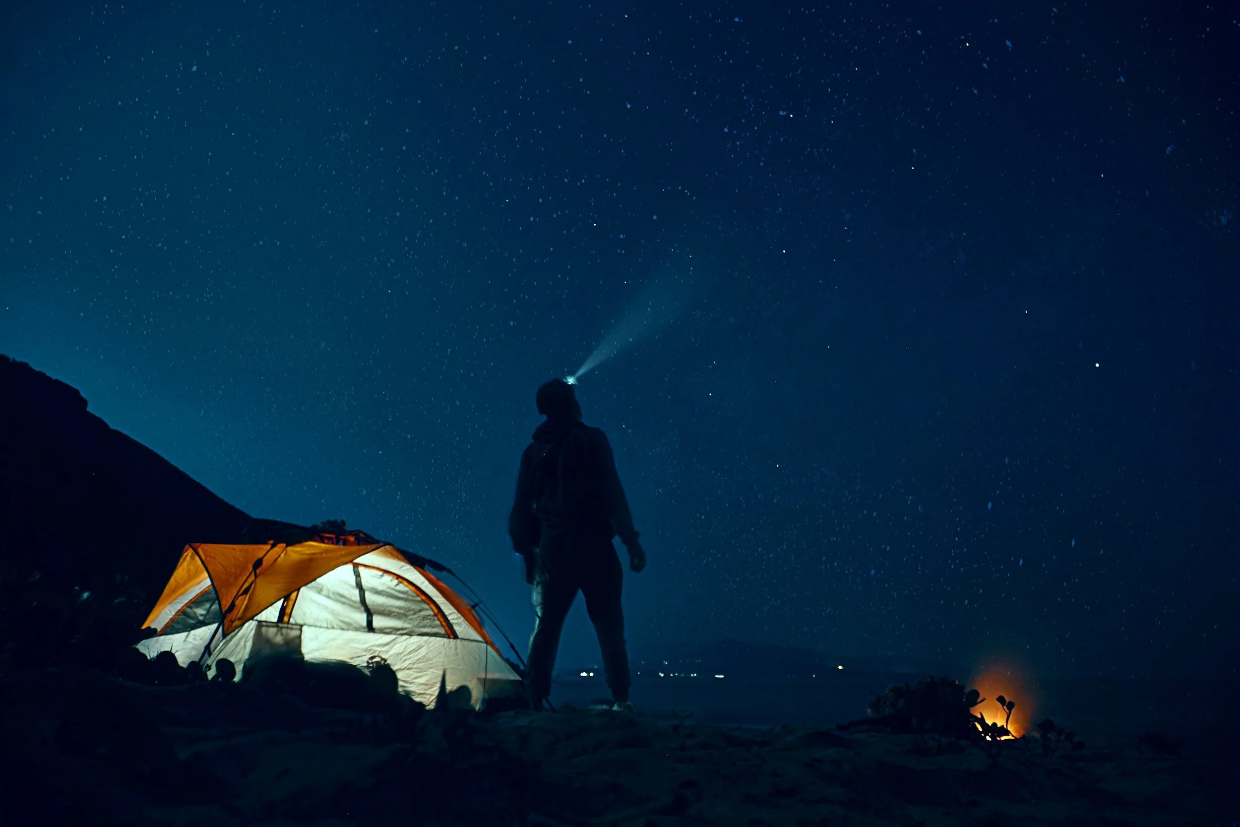 A person standing on a beach at night near a tent, looking up at a star-filled sky with a flashlight attached to their forehead.