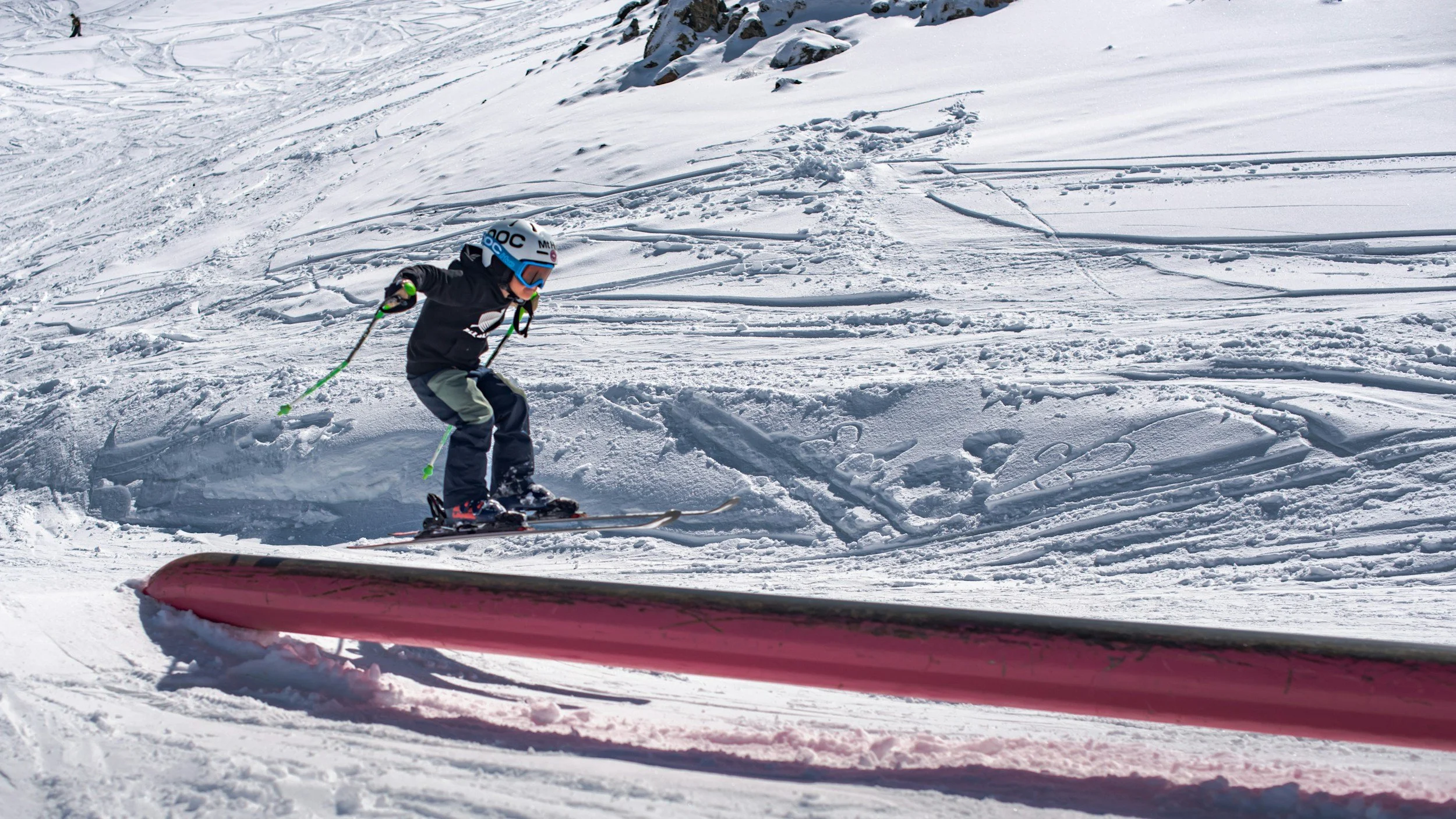 Young child skiing downhill on a snowy slope, wearing a helmet and goggles, with ski poles in hand.