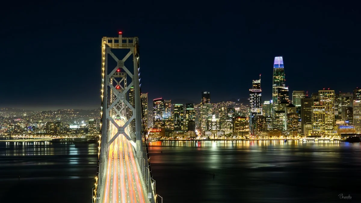 Night view of the San Francisco skyline with tall illuminated buildings and a lit-up suspension bay bridge over water.