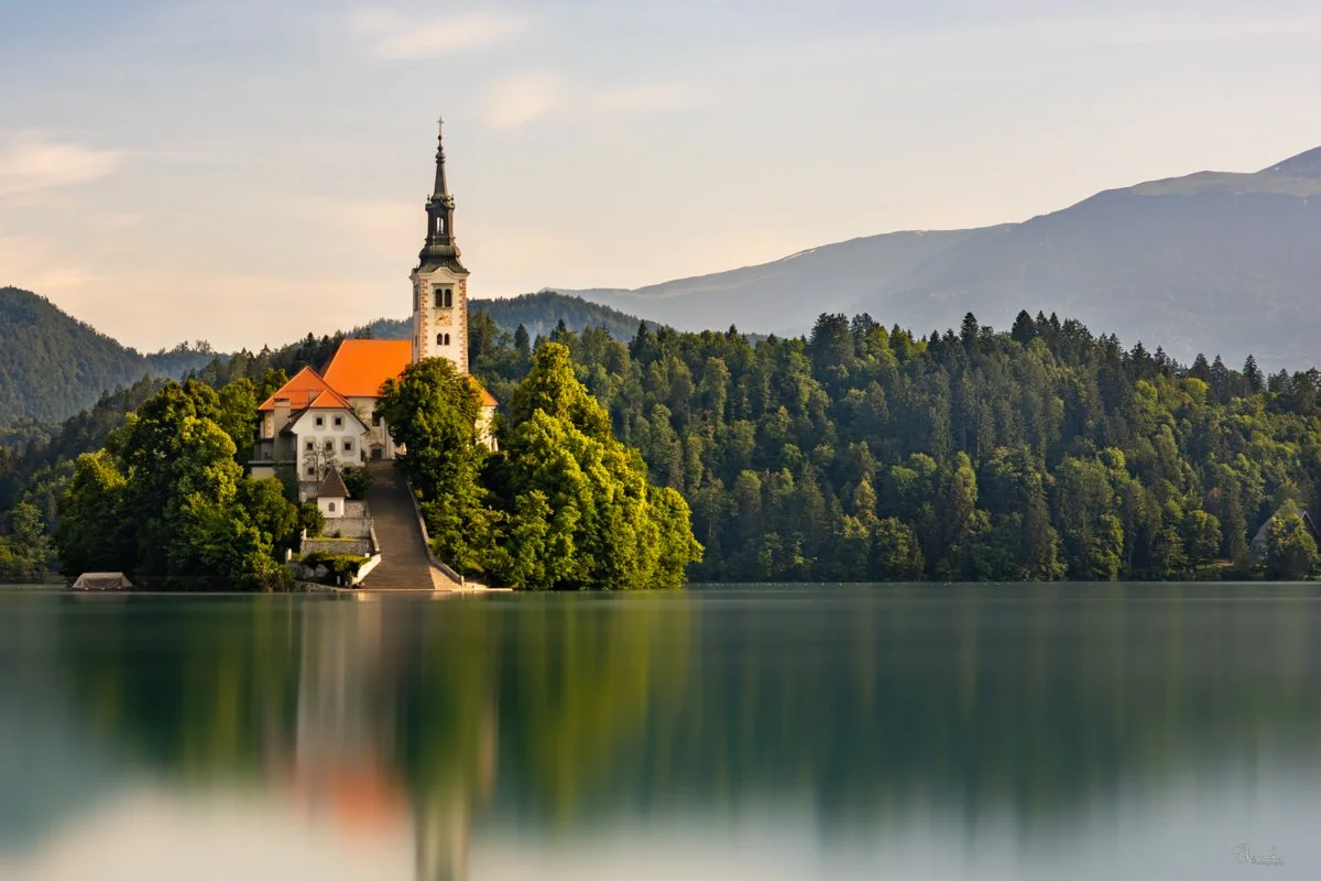 The church in Lake Bled with a tall steeple on a small, forested hillside island surrounded by a calm lake, with mountains in the background.