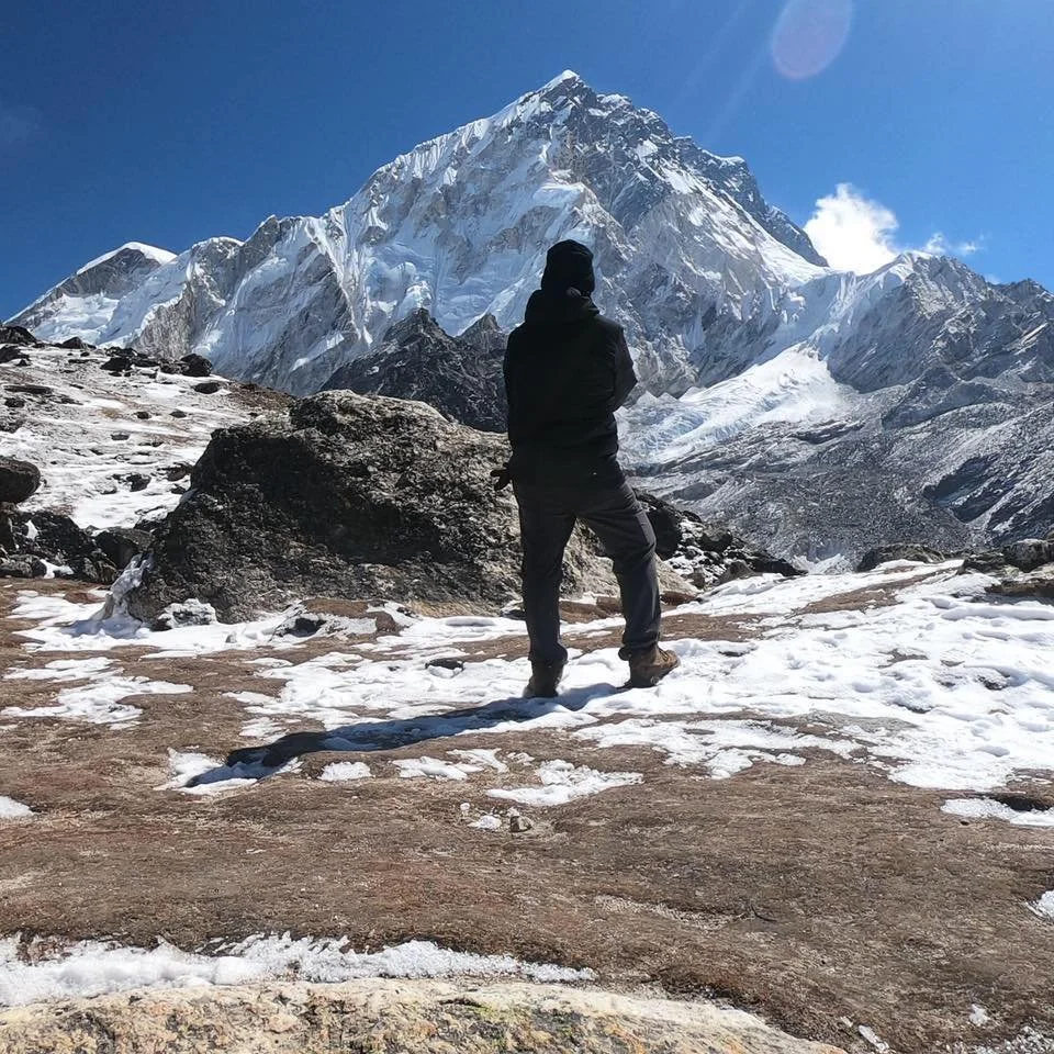 A person dressed in black standing on snow-covered ground, looking at a large snow-capped mountain under a clear blue sky.