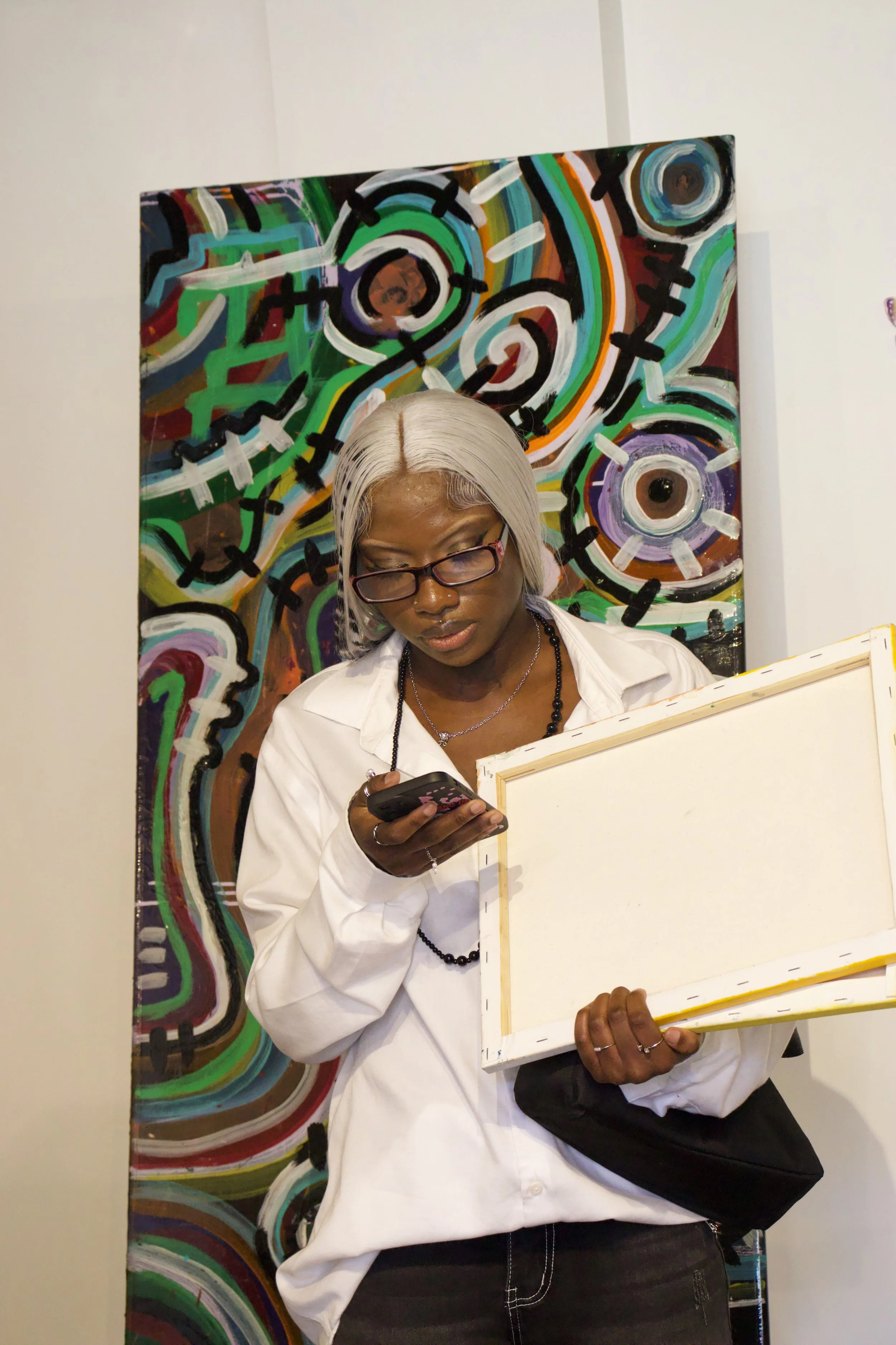 A woman with silver hair, wearing glasses, a white shirt, a black beaded necklace, and holding a phone and an artist's palette, standing in front of a colorful abstract painting.