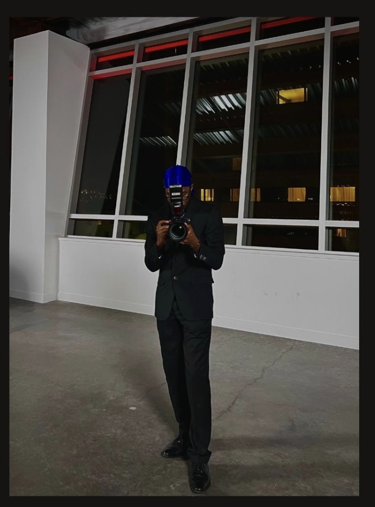 A person in a black suit and blue helmet taking a photo with a camera in front of large windows at night.