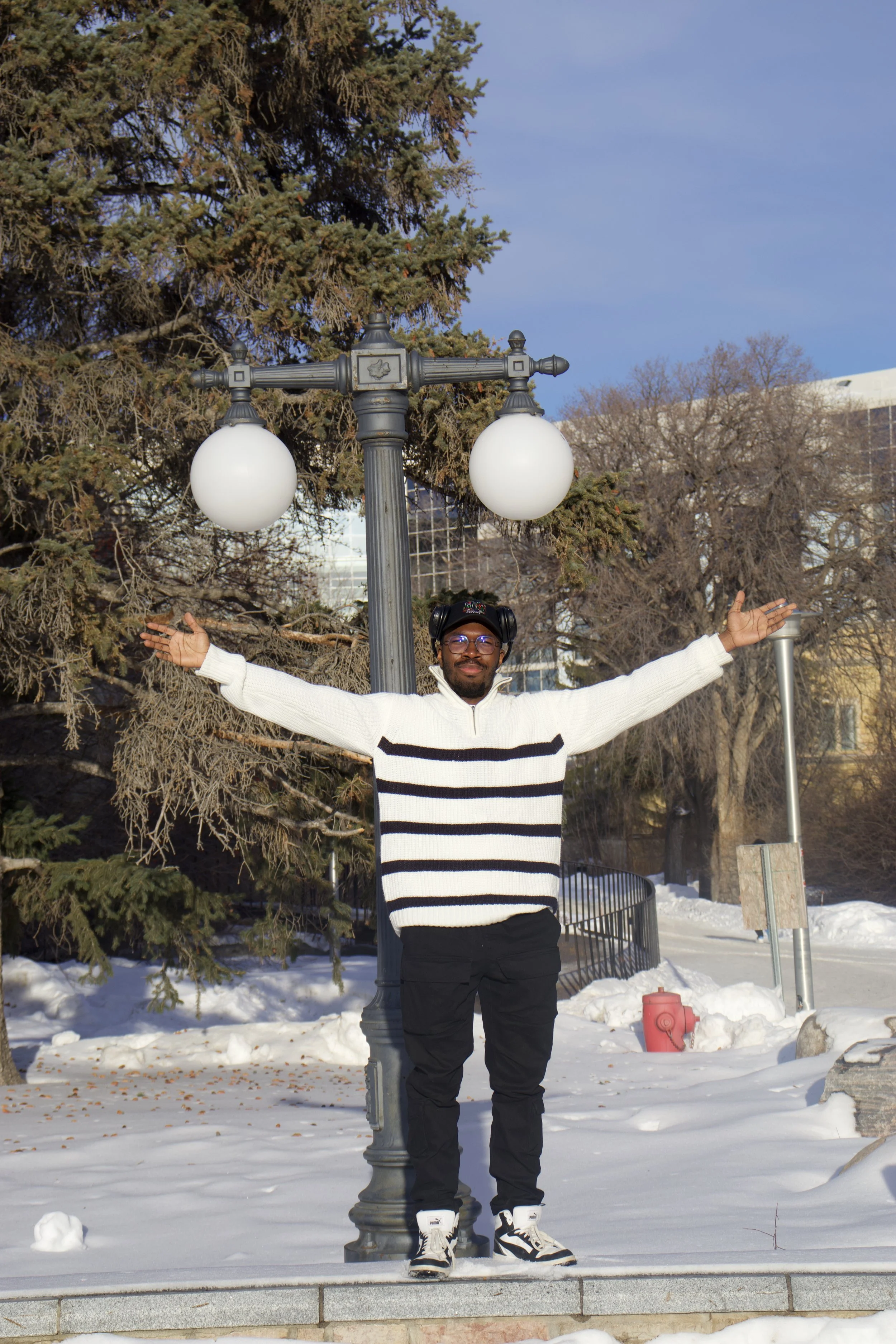 A man stands outdoors in winter snow with arms outstretched, smiling, in front of a lamppost with two globe lights, trees, and buildings in the background.