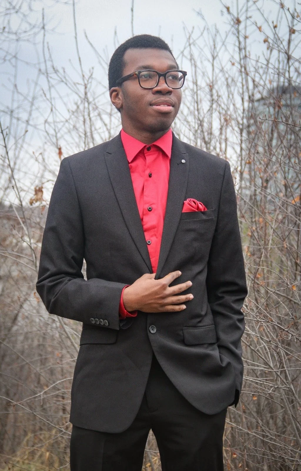 A young man in a black suit with a red shirt, red pocket square, and glasses, standing outdoors in front of leafless trees.