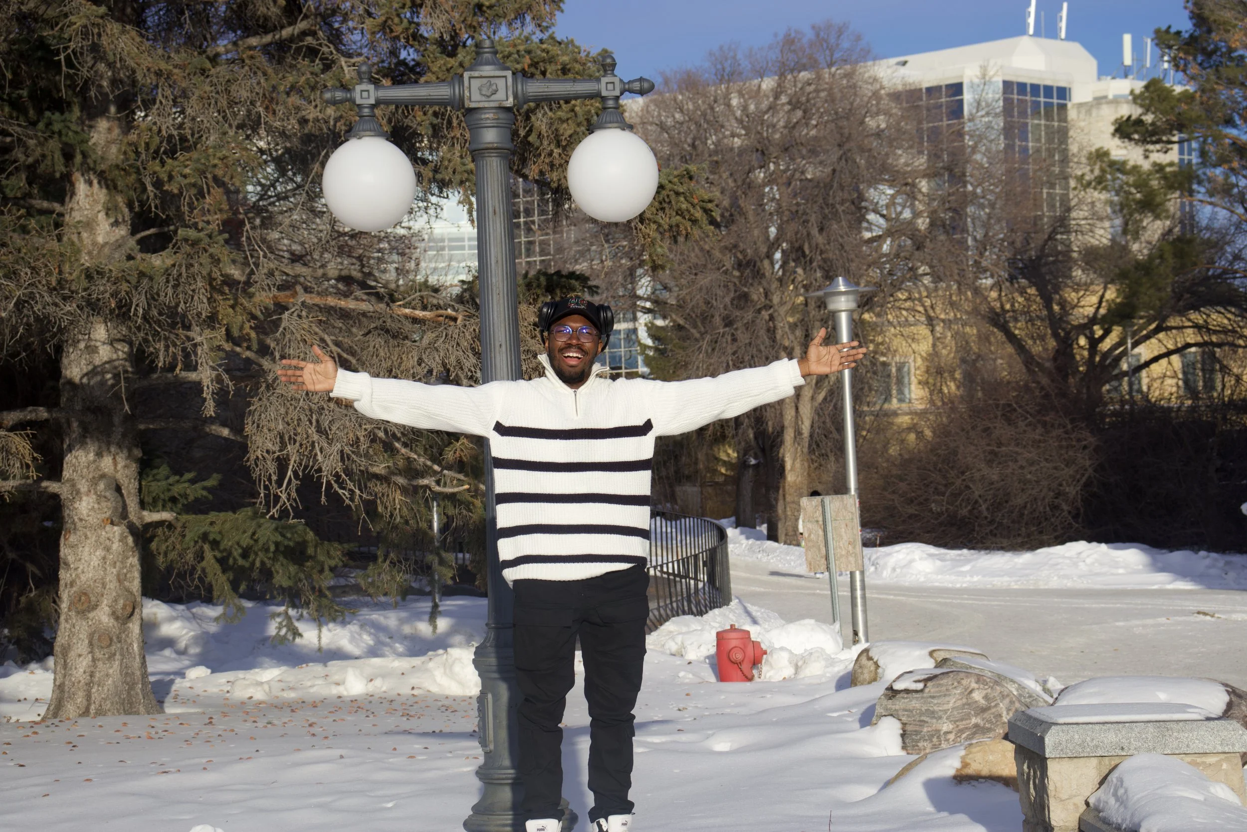 A joyful man standing outdoors in snowy park, arms outstretched, smiling, with lamp posts and trees in background.