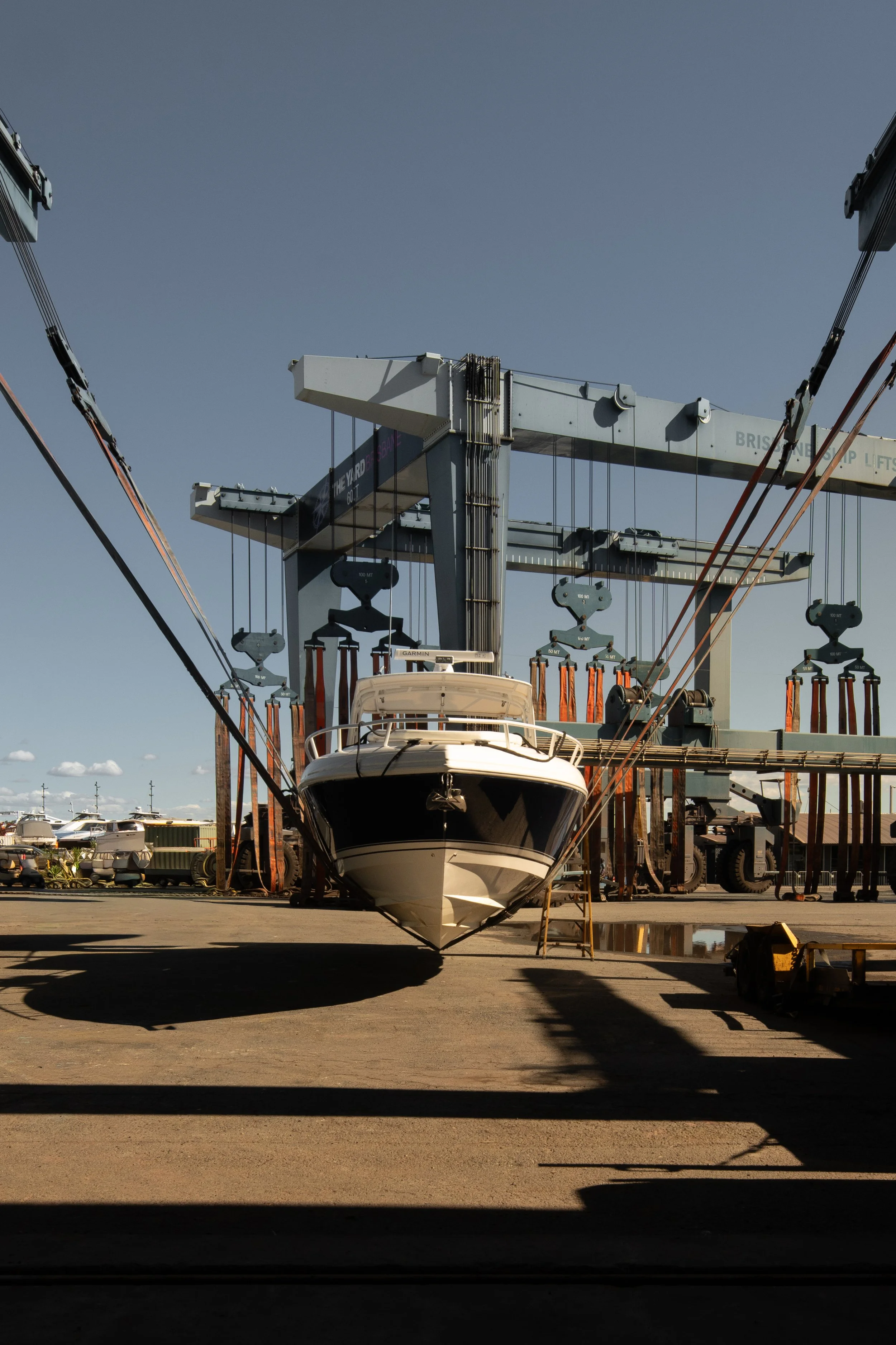 A boat is suspended in a boatyard by a large crane, with other boats and equipment visible in the background under a clear blue sky.