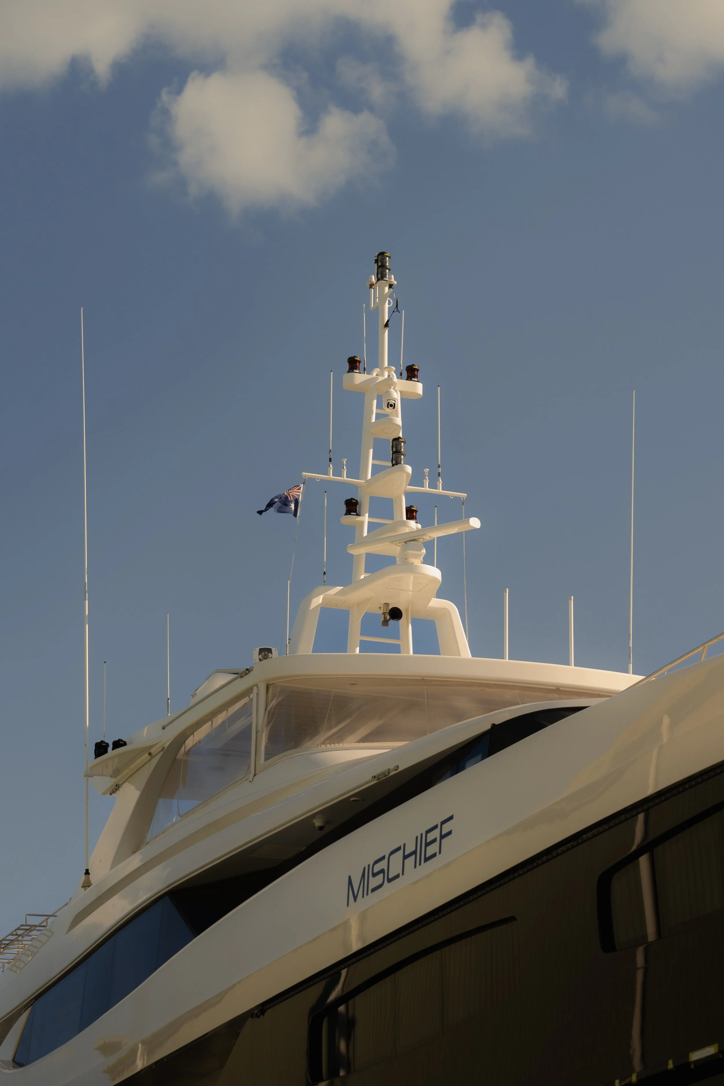 Close-up of a luxury boat's upper deck with antennas, navigation equipment, and the name 'MISCHIEF' on the side, set against a blue sky with scattered clouds.
