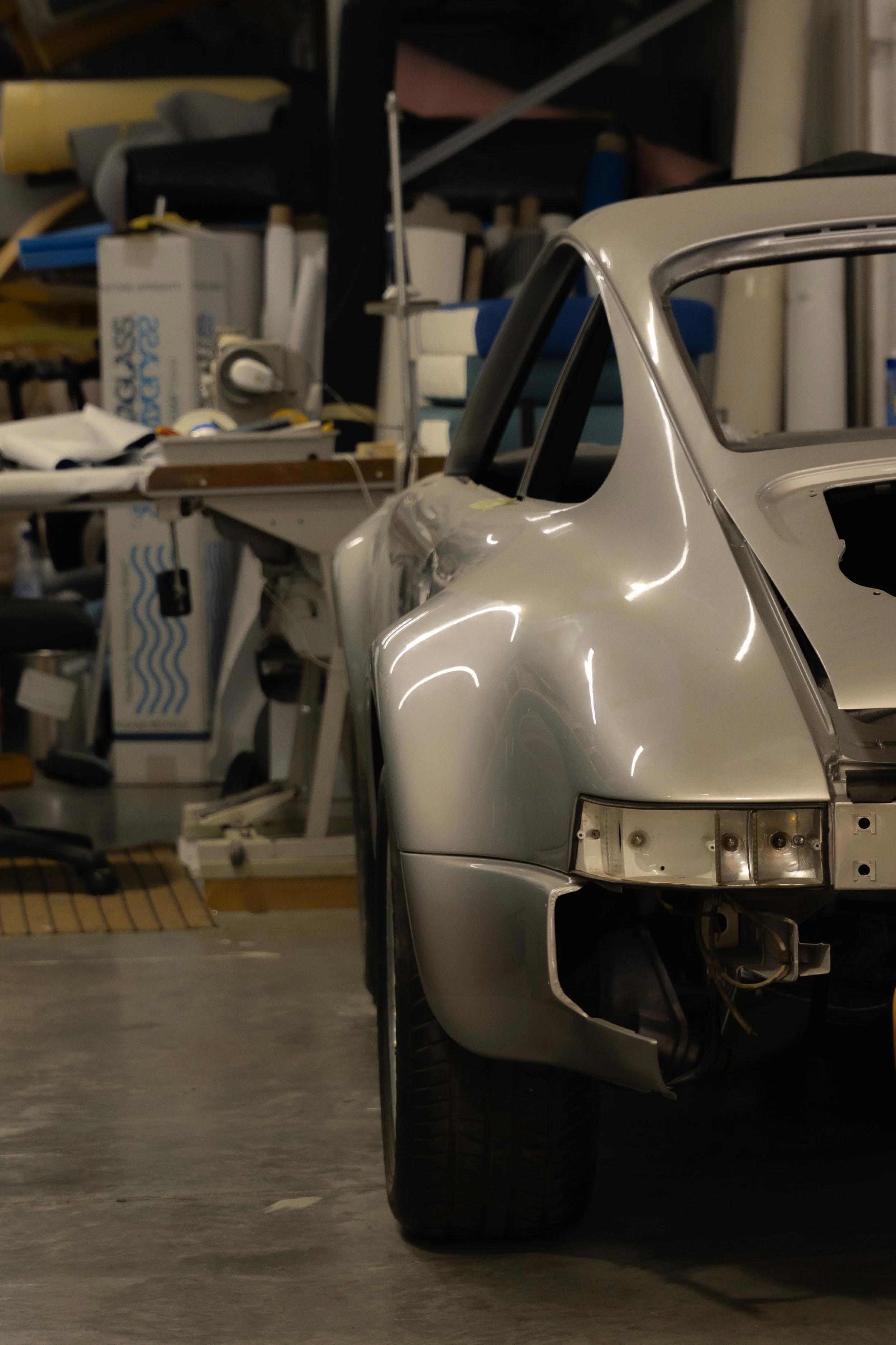 Partially assembled silver sports car in a garage. The car's front section, including the wheel and headlight area, is visible. Background contains shelves cluttered with boxes, tools, and equipment.