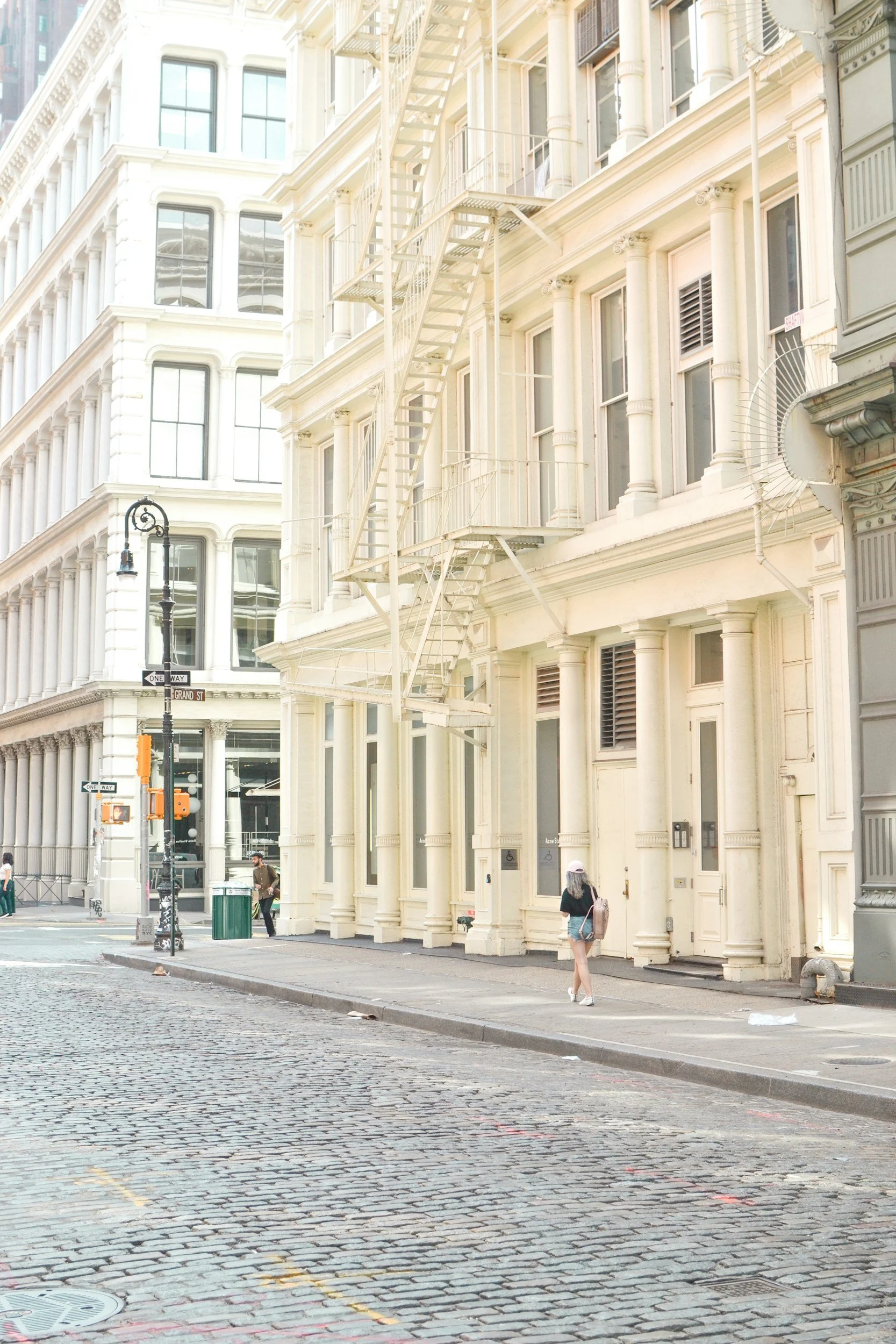 Street view of cream-colored historic buildings with fire escapes, sidewalk, and pedestrians, in an urban city neighborhood.