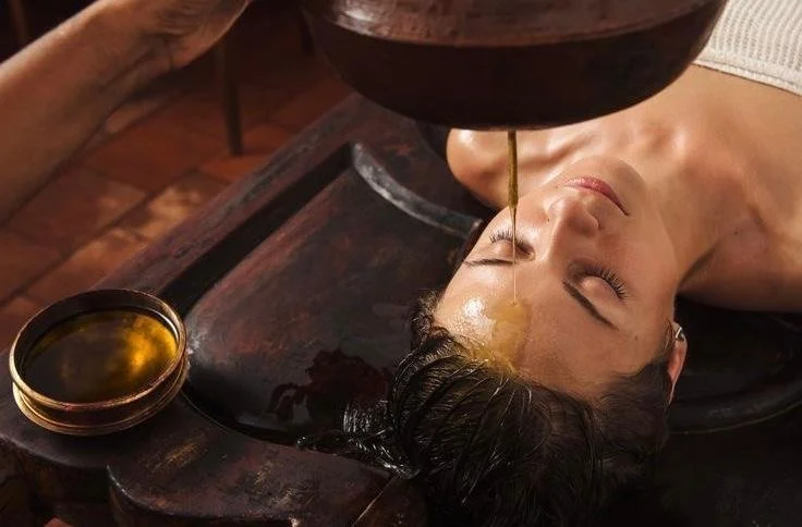 A woman receiving a facial treatment with a thick liquid or oil being poured onto her forehead while she relaxes with her eyes closed.