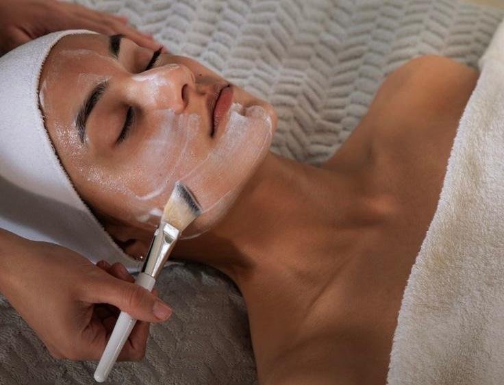 A woman receiving a facial treatment, lying down with a towel wrapped around her head, her eyes closed as a skincare professional applies a white facial mask with a brush.